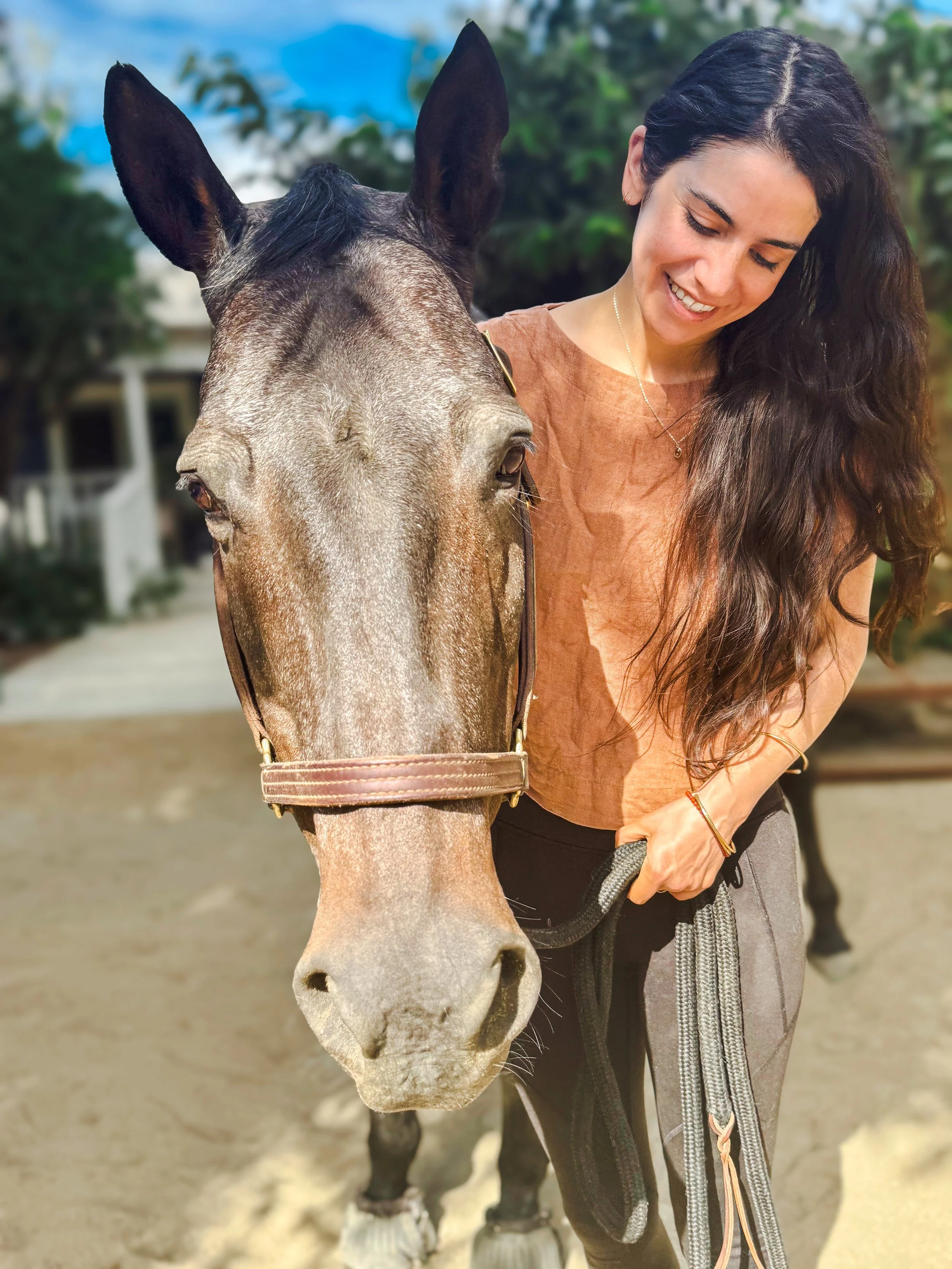 A woman standing next to a brown horse outdoors, smiling and holding its reins.
