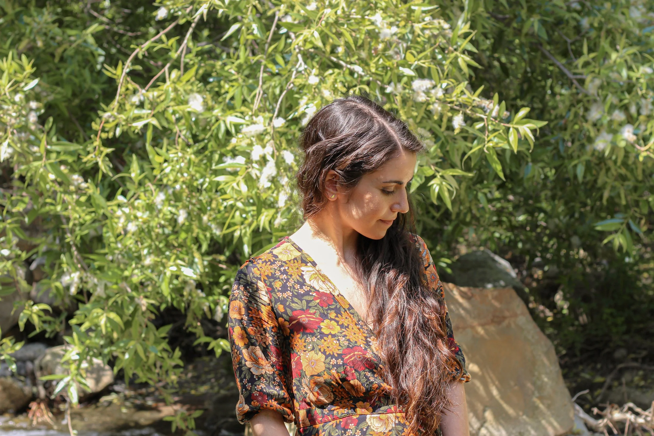 A woman with long wavy brown hair wearing a floral dress looks down outdoors surrounded by green foliage and rocks.