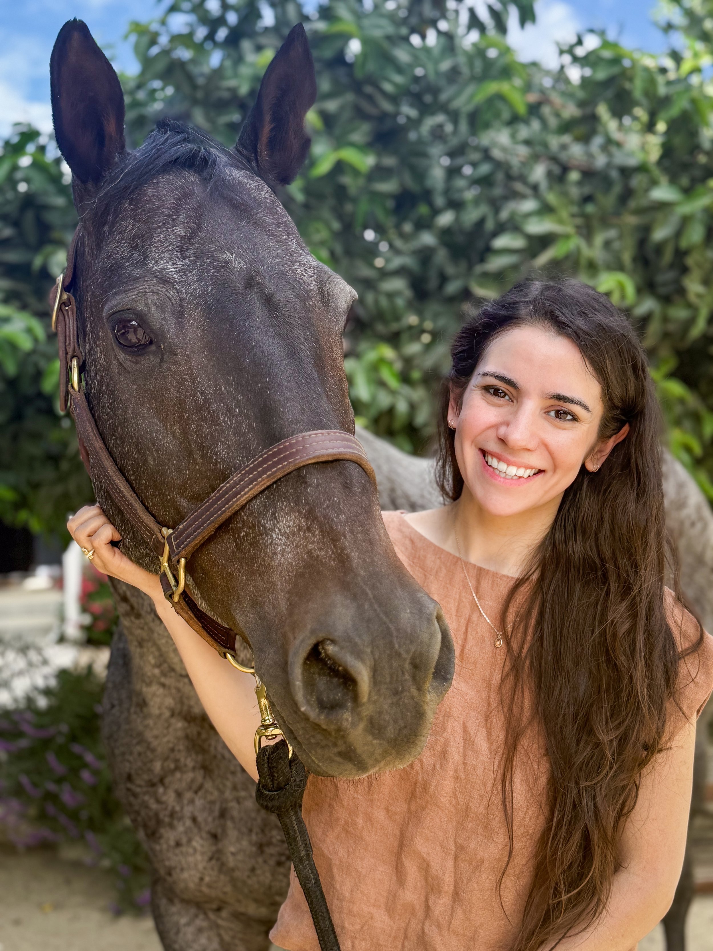 A young woman with long brown hair smiling and holding a brown horse by the halter in an outdoor setting with trees and greenery.