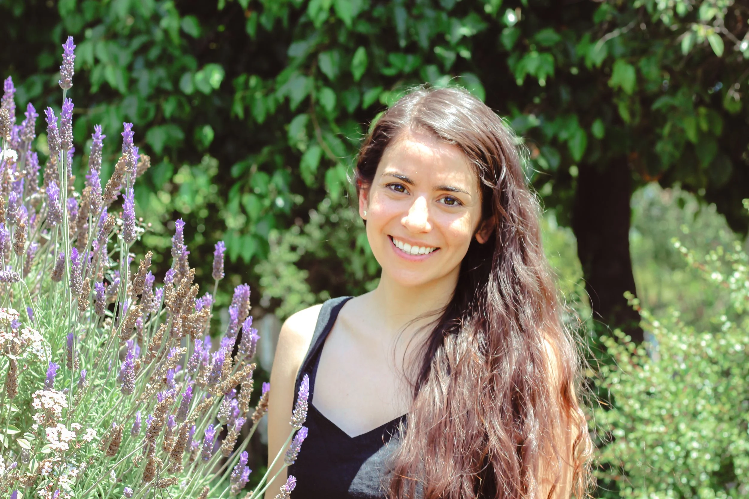 A young woman with long wavy brown hair and a black sleeveless top standing outdoors near purple lavender flowers, with green trees in the background, smiling at the camera.
