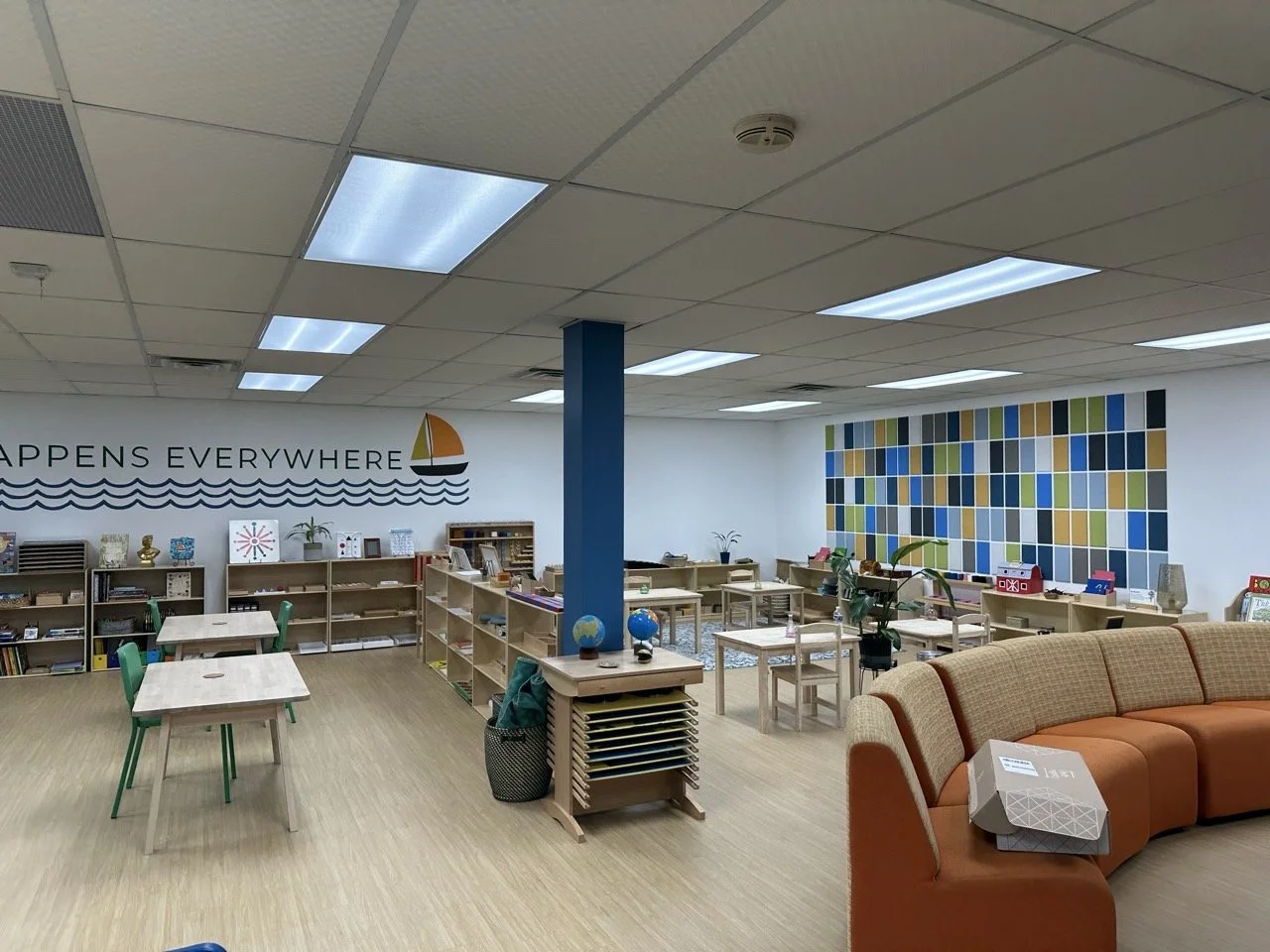 A spacious preschool classroom with small wooden tables and green chairs. There are educational materials, books, and plants on the shelves and tables. Large windows let in natural light, and a colorful flag banner decorates the room.