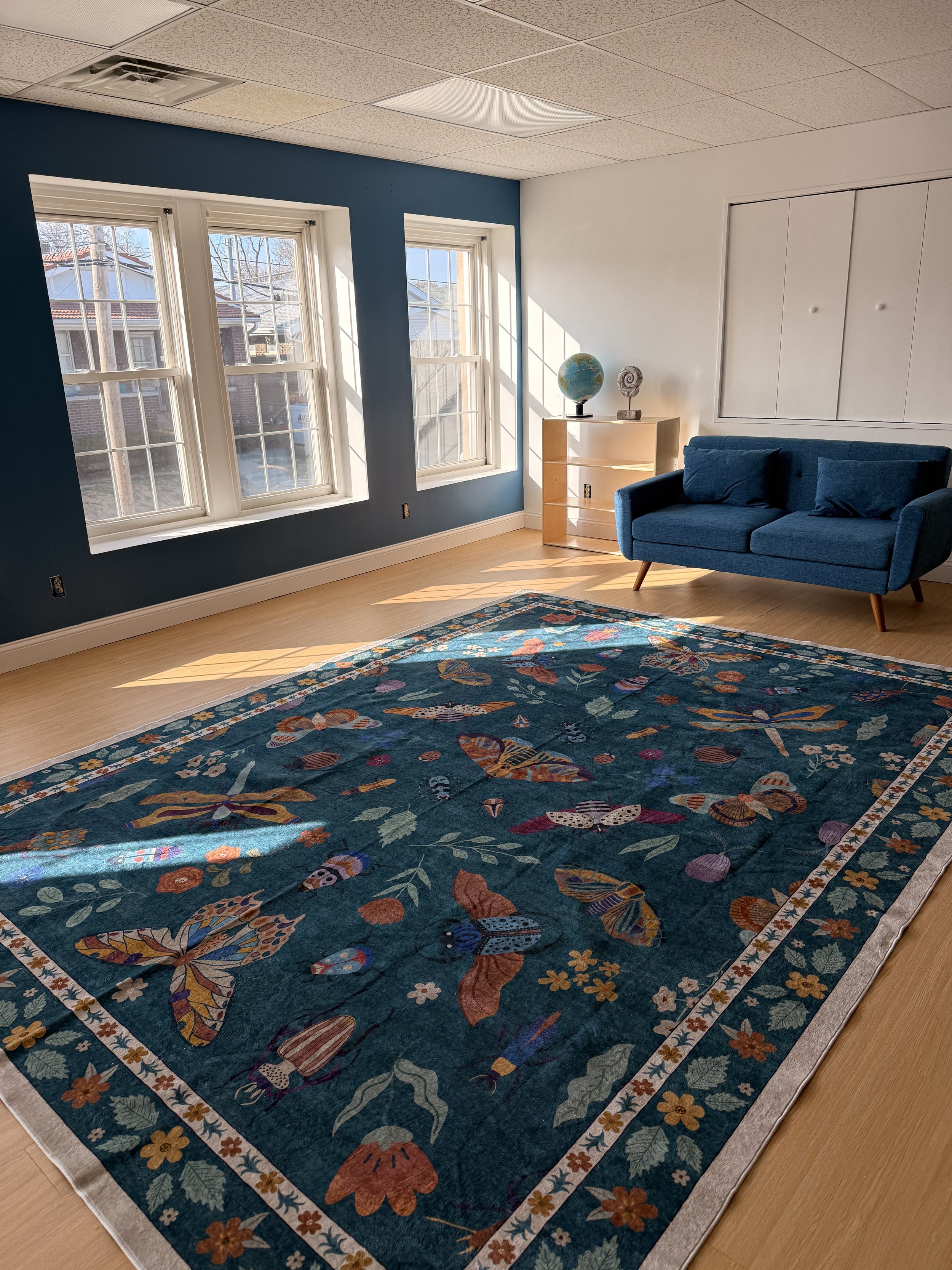 A bright room with three large windows, a blue sofa, a wooden shelf with a globe and decorative item, and a colorful butterfly-patterned area rug on light-colored wooden floors.