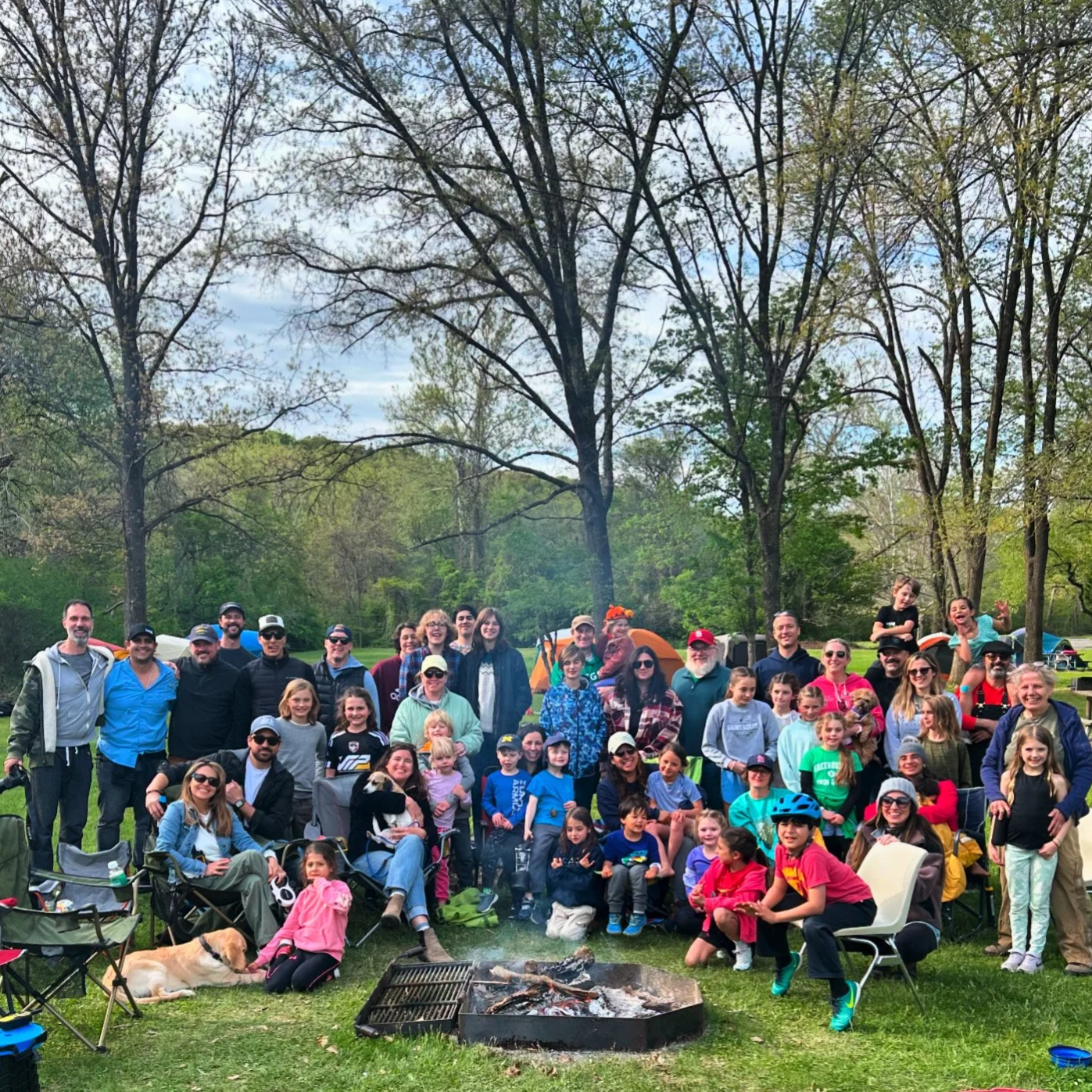 A large outdoor gathering of people, children, and dogs in a park setting during daytime. The group is around a campfire with tents and trees in the background.
