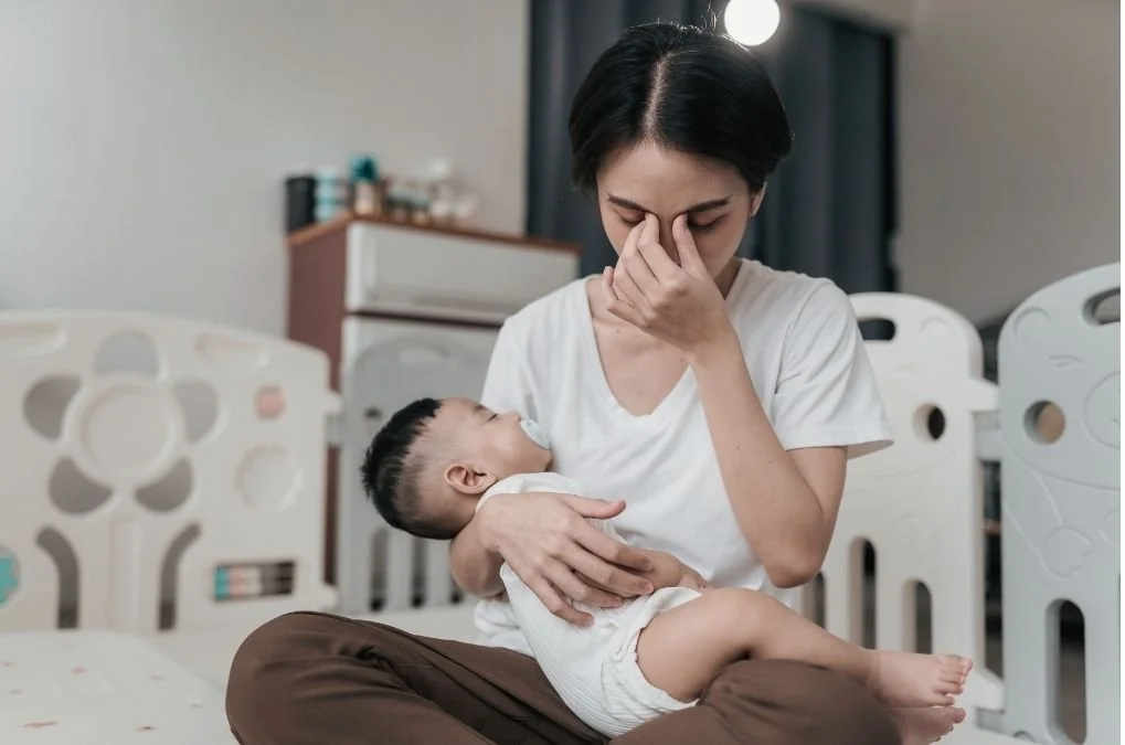 Mother holding her baby, rubbing her eyes looking tired.