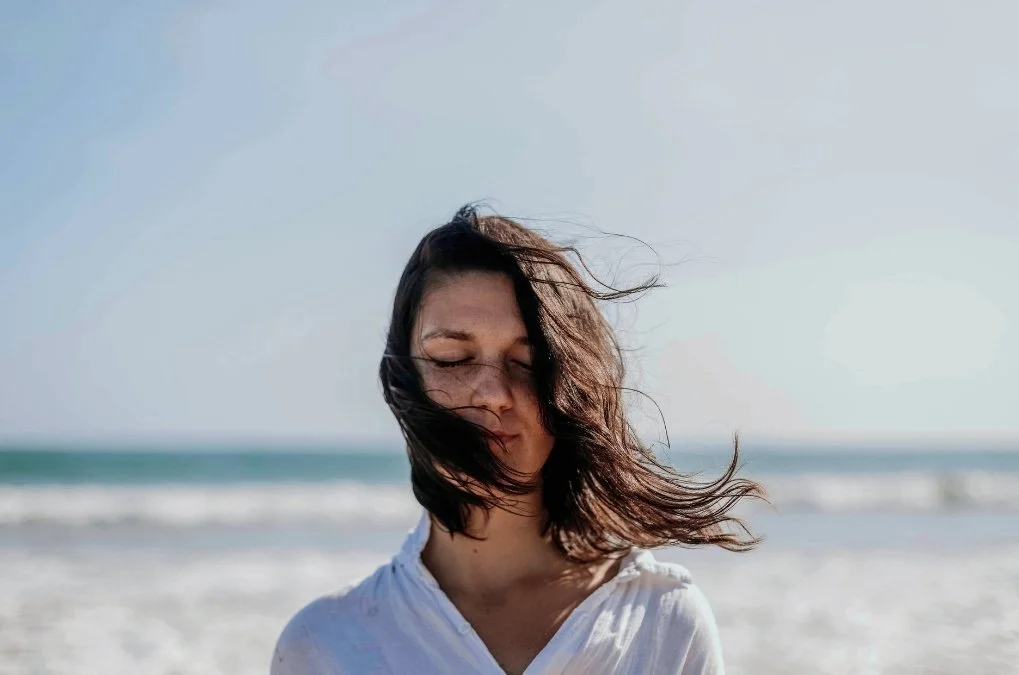 Soft abstract background of woman standing at the beach with eyes closed, hair blowing in the wind. for the section: What does intuitive eating therapy look like at MLP therapy group?