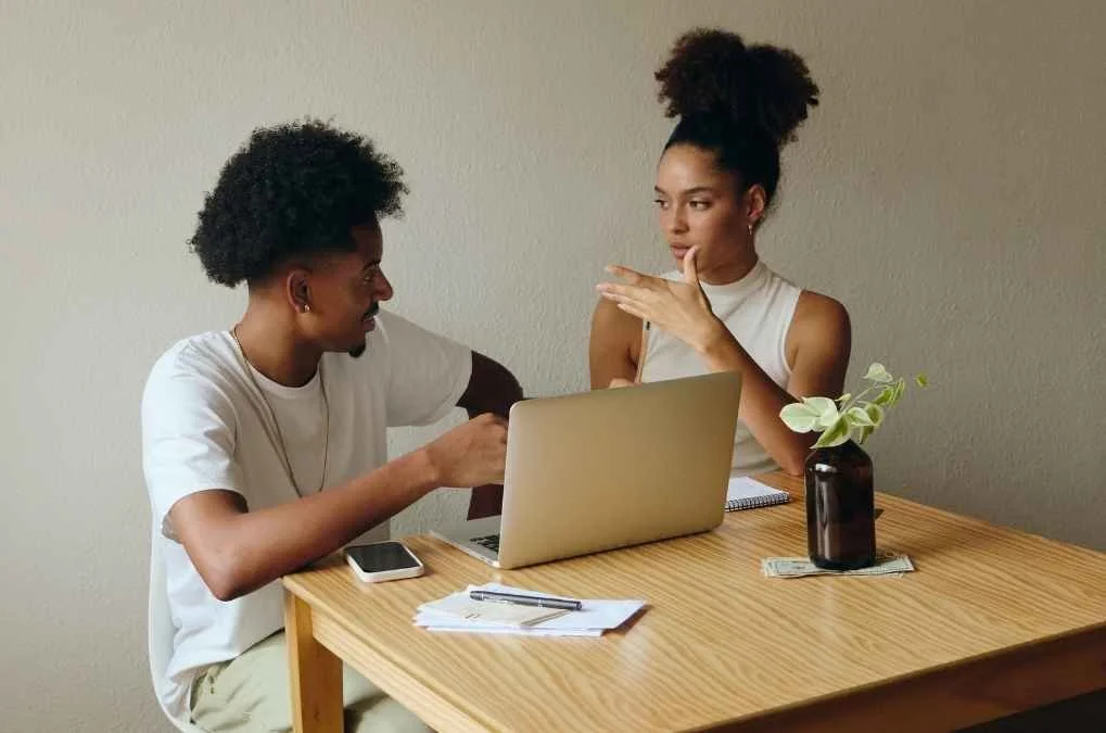 Couple sitting at a table looking at a laptop, mid-conversation.