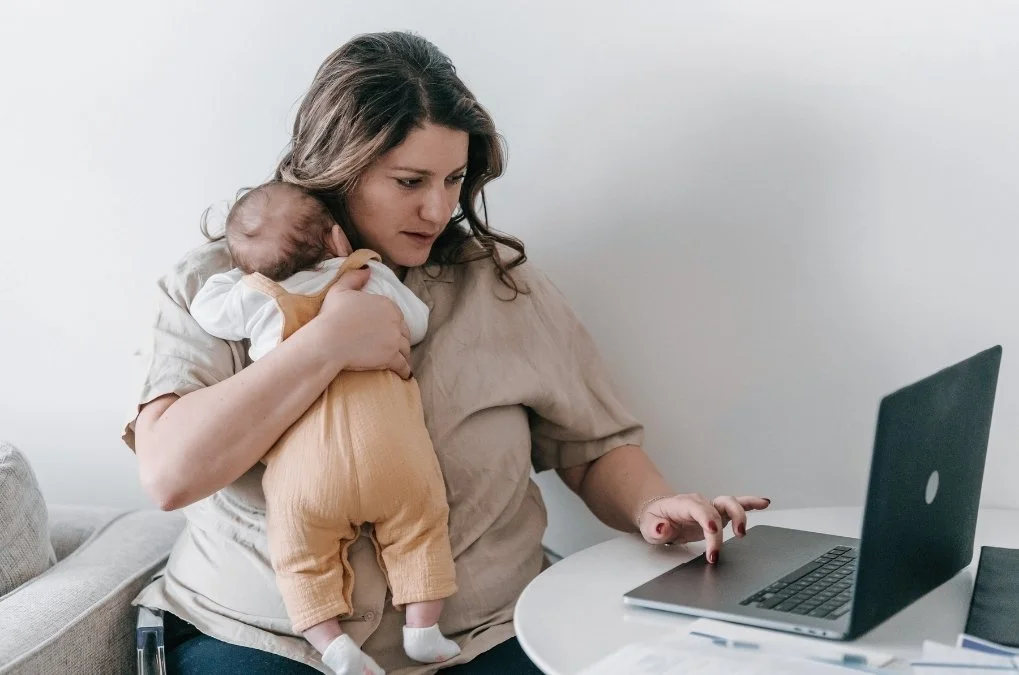 Mother holding a baby while working on her laptop.