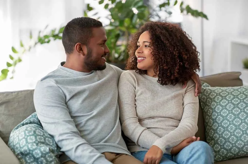 Couple smiling at each other on a couch, arm around her shoulder.