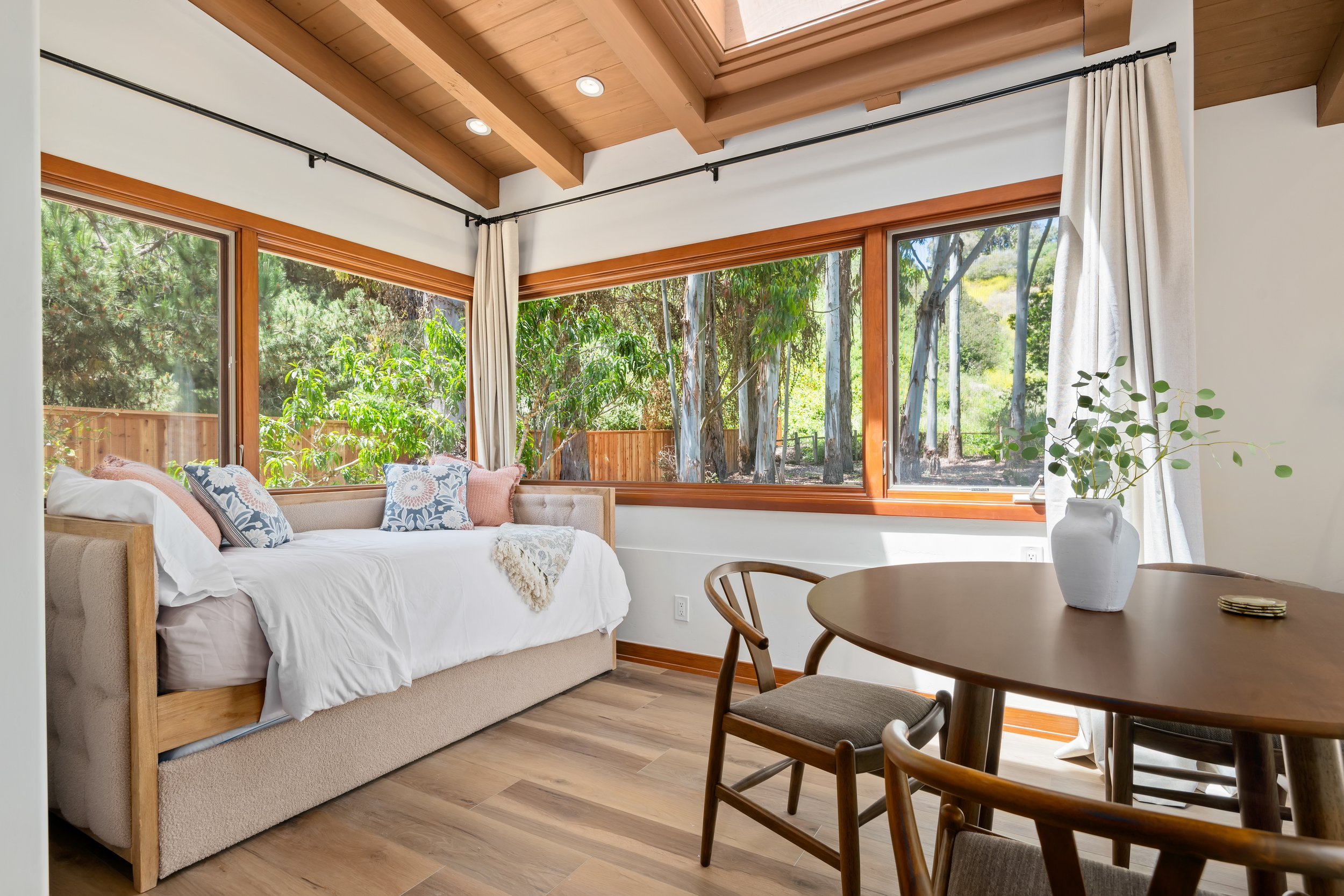 Living room with large windows showing outdoor trees, a white sofa with decorative pillows, a round wooden dining table with chairs, and a ceramic vase with green foliage.