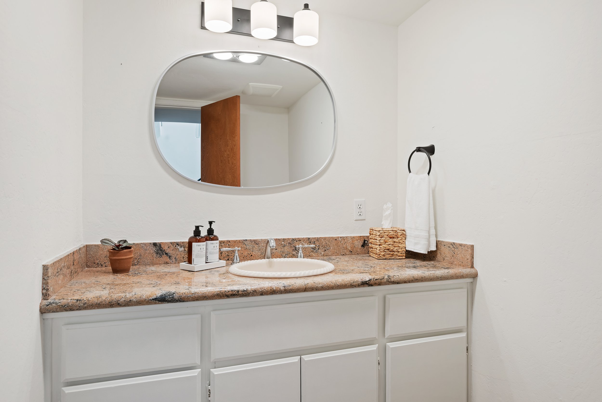 Bathroom vanity with a granite countertop, white cabinets, a round sink, a large oval mirror, and small decorative items including lotion bottles, a small potted plant, a woven tissue box, and a hand towel on a black metal towel ring.