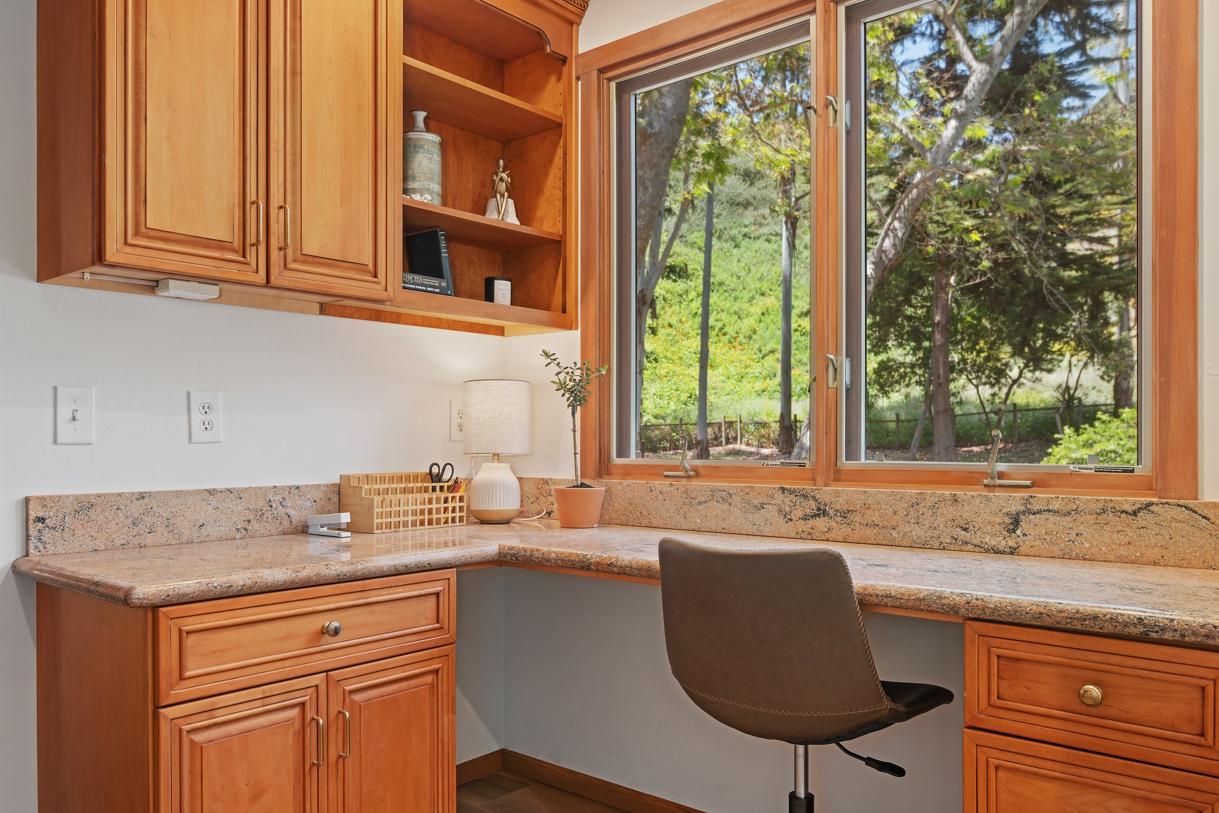 Home office workspace with wooden cabinetry, granite countertop, a potted plant, a desk lamp, and a view of trees through large windows.