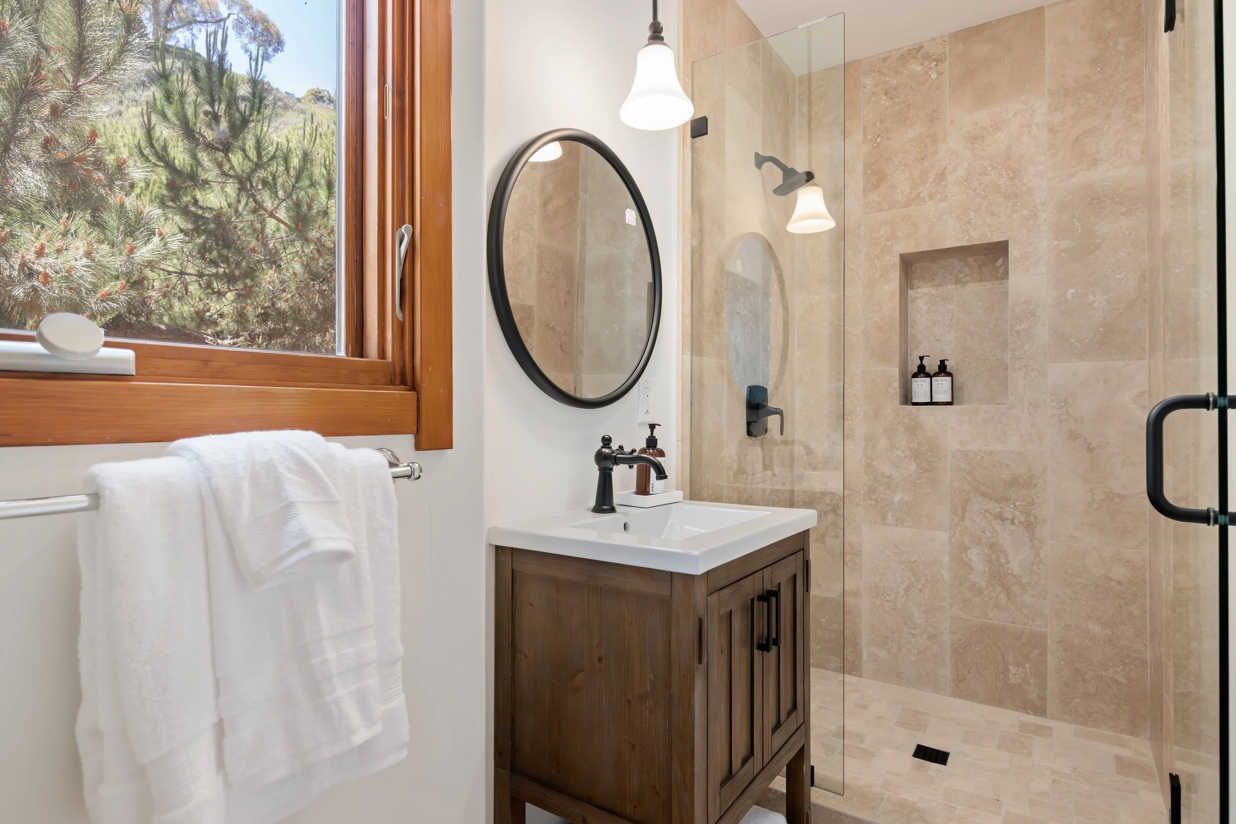 Bathroom with a wooden-framed window showing trees outside, a white towel hanging on a rack, a small wooden vanity with a white sink and a black faucet, an oval mirror, and a walk-in shower with beige tiles, a built-in niche holding bottles, and mode