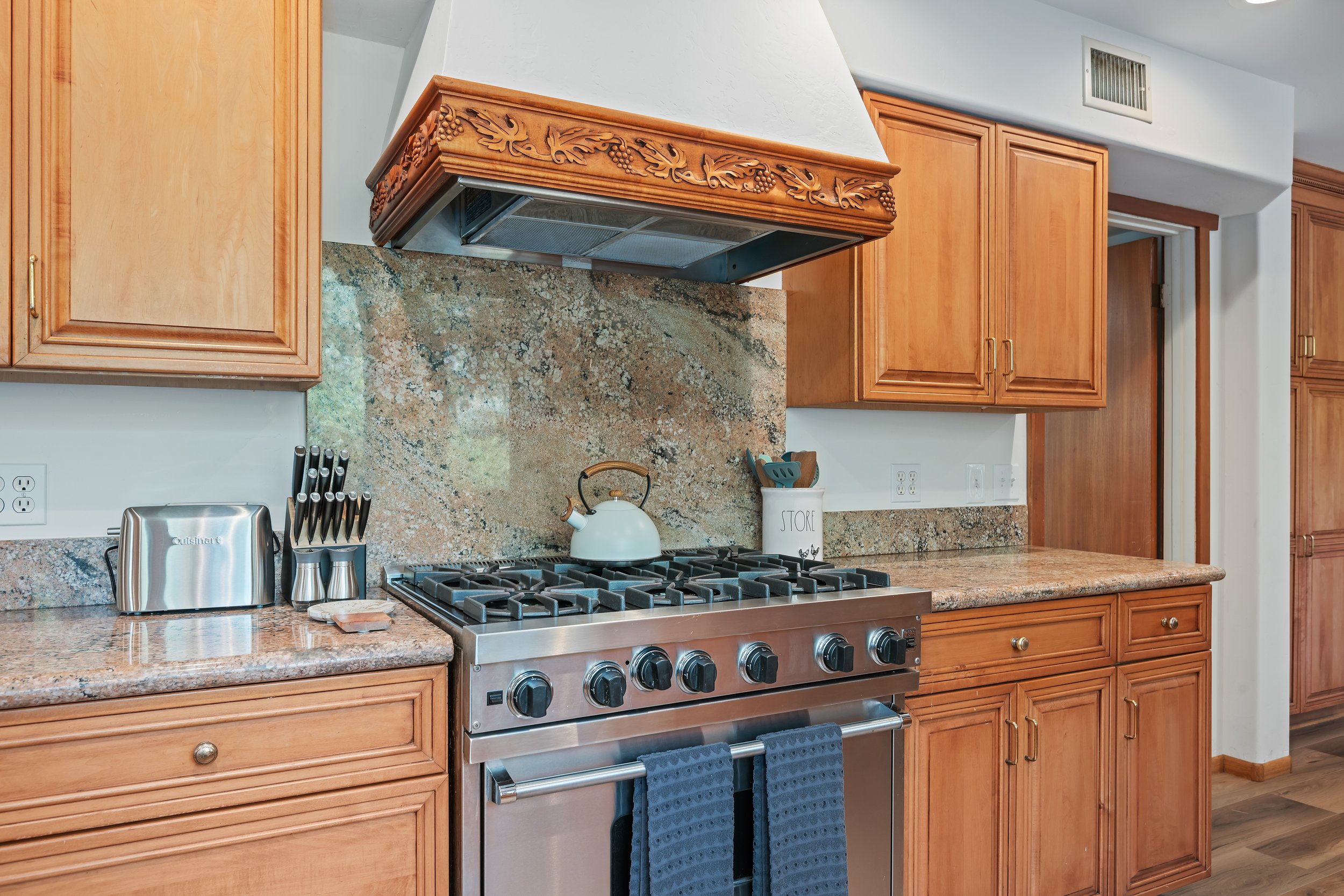 Kitchen with wooden cabinets, granite countertops, and a stainless steel stove with a white teapot on top.