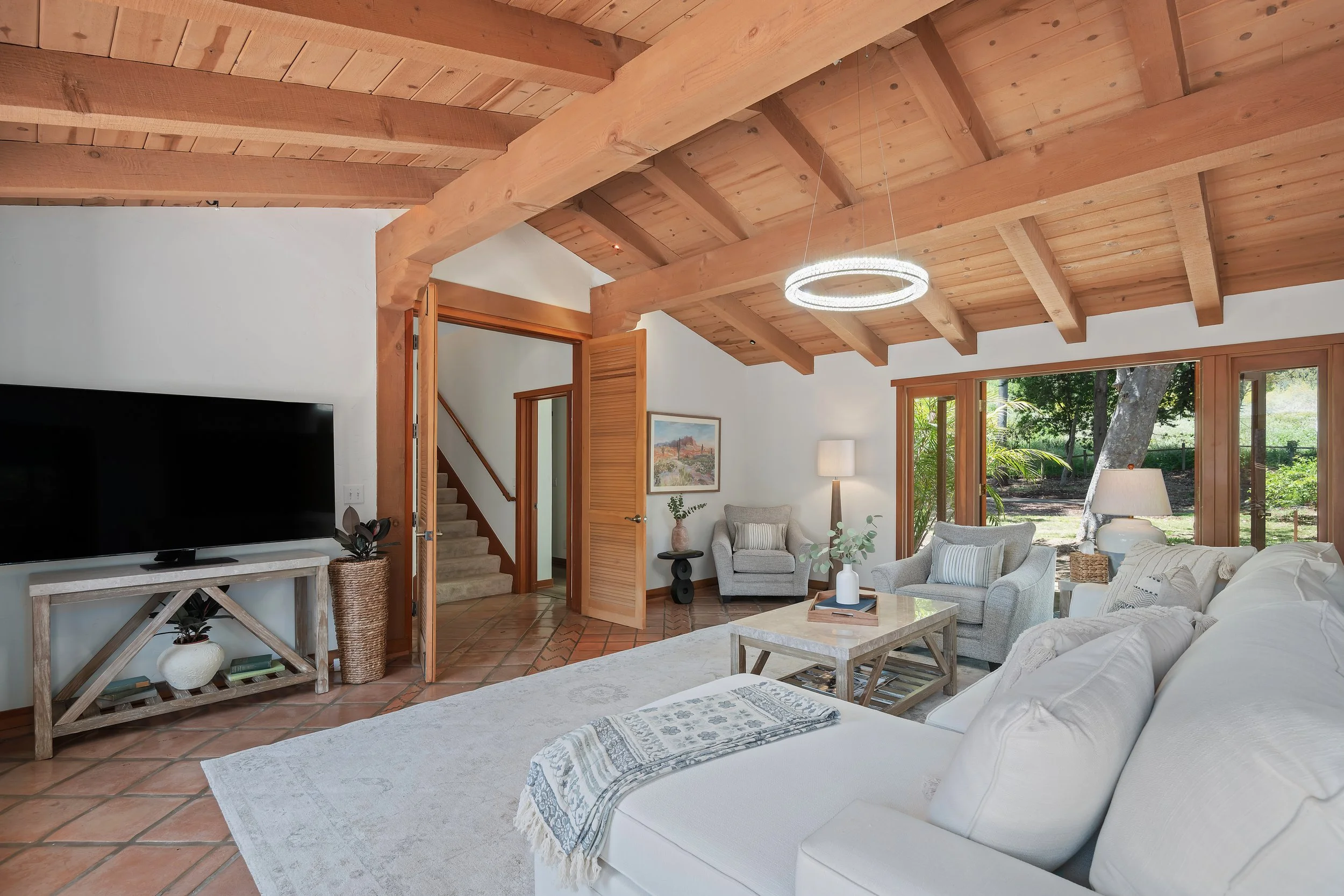 Living room with white sofa, armchairs, wooden ceiling beams, large windows showing outdoor trees, and a flat-screen TV on a rustic console.