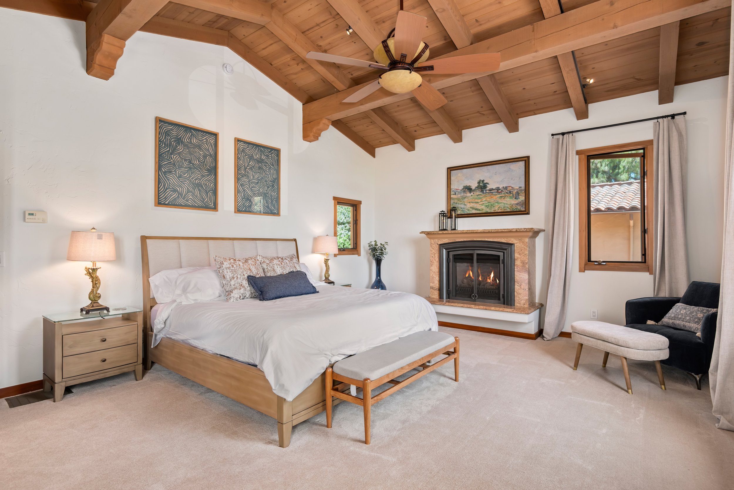 A bedroom with a wooden ceiling, a bed with white bedding and patterned pillows, two bedside lamps, framed artwork above the bed, a fireplace with a painting above, a window with curtains on the right, and a black armchair with an ottoman.