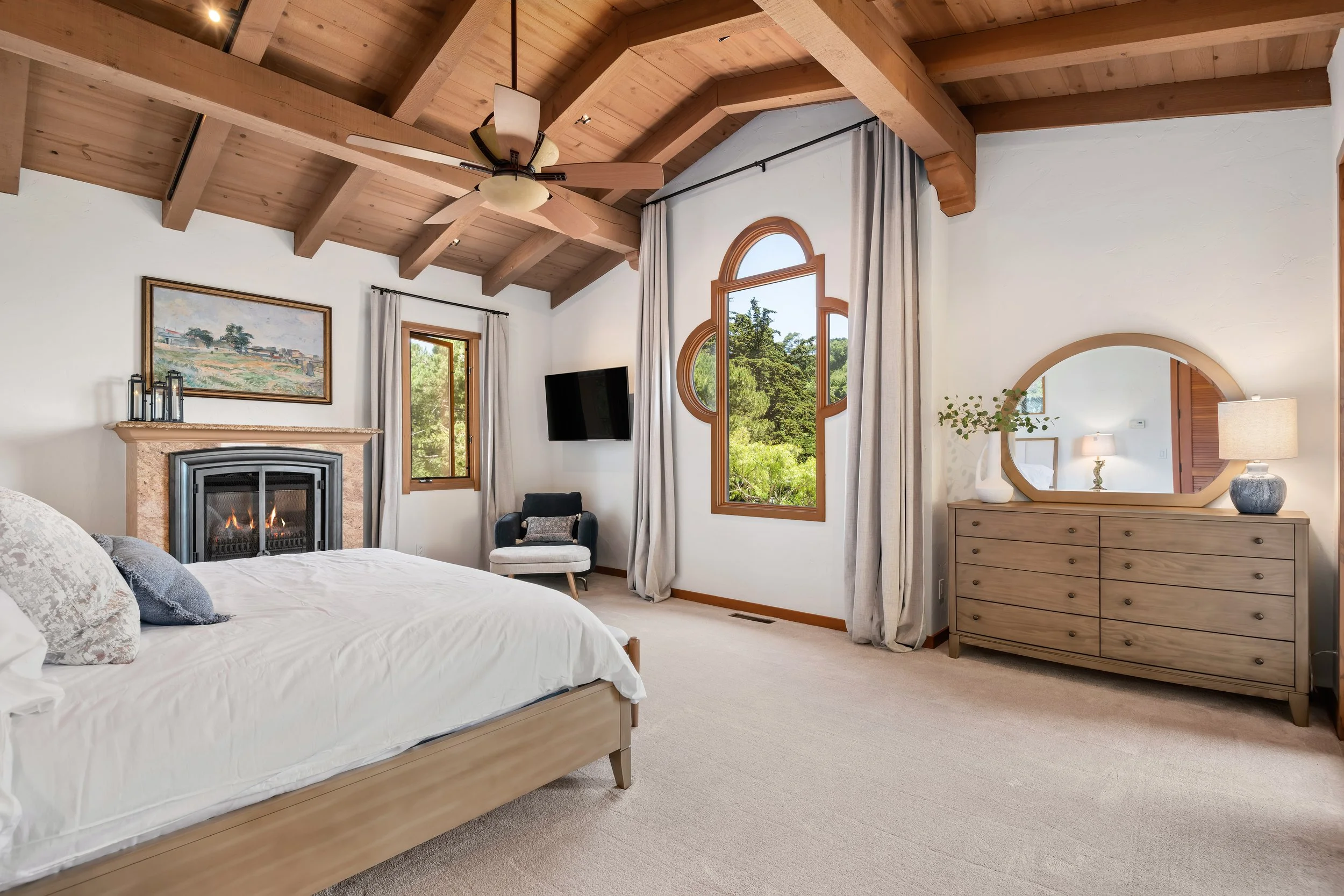 Bedroom with a wooden vaulted ceiling, a bed with white bedding, a fireplace with artwork above, window and curtain, armchair, wall-mounted TV, dresser with mirror, and large windows showing greenery outside.