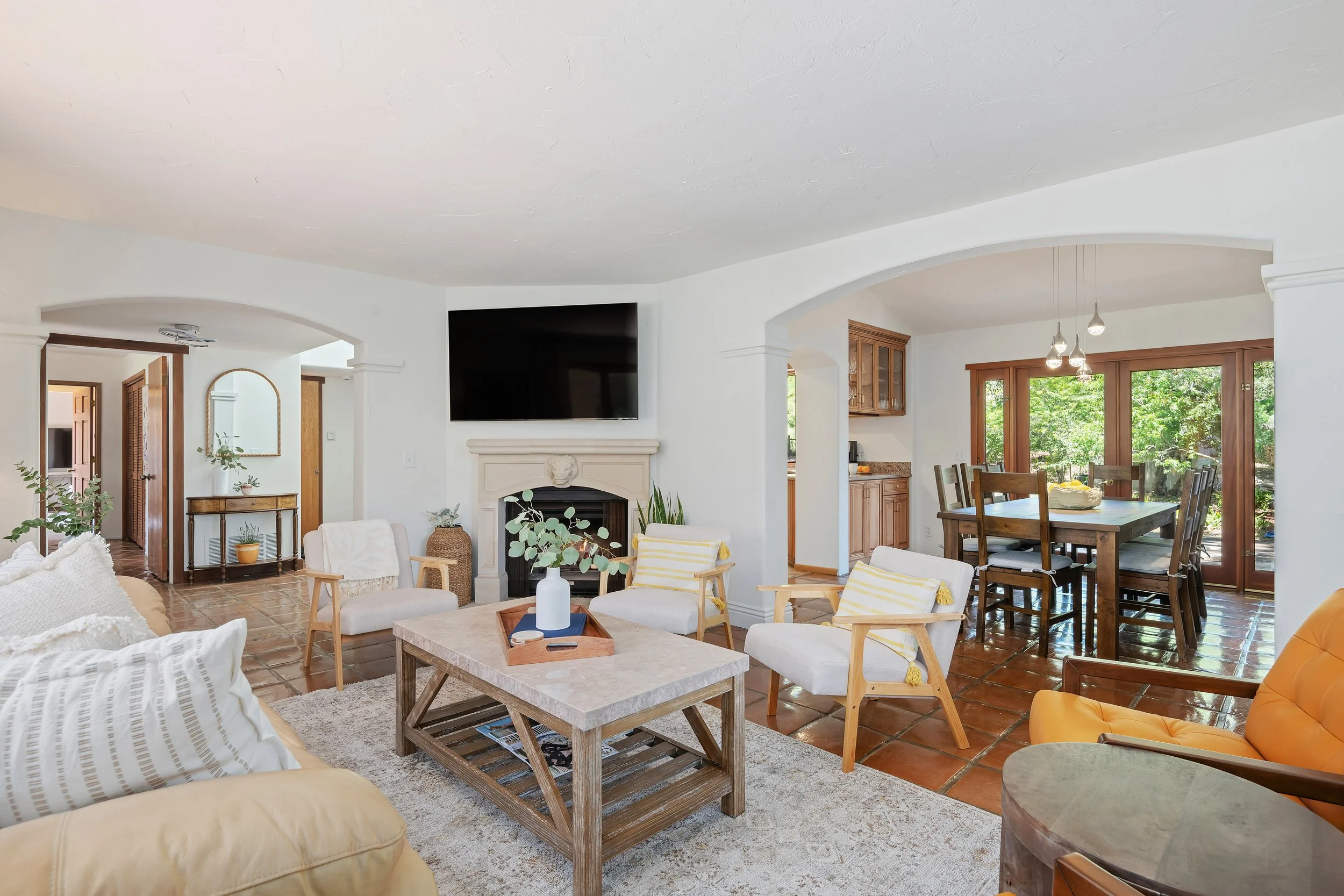 Living room with white walls, wooden floor, a fireplace with a TV above, beige sofas, armchairs, a wooden coffee table, and large glass sliding doors with a view of trees outside.