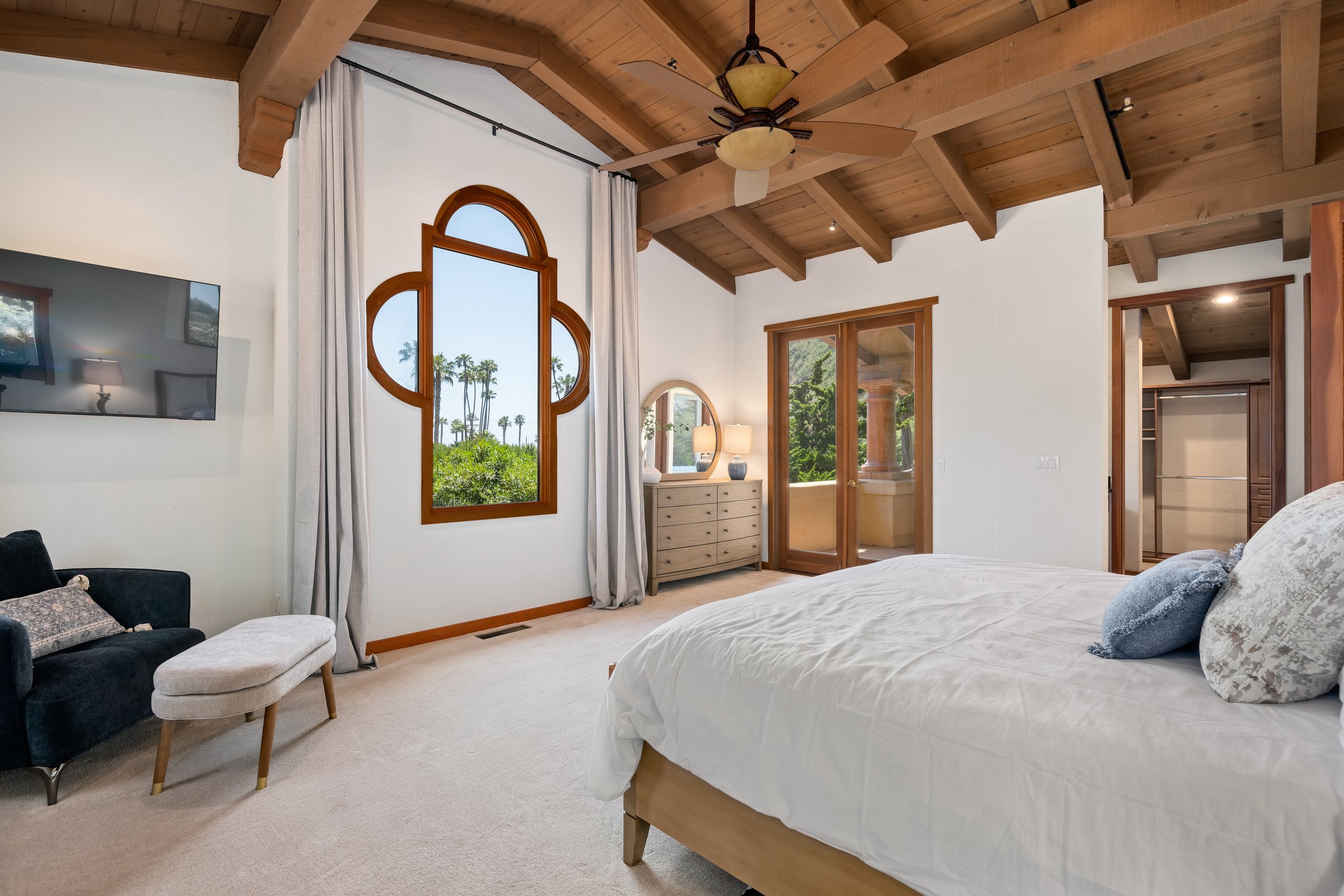 A spacious bedroom with a wooden ceiling with beams, a large bed with white bedding, a dresser with a mirror, a small table with a lamp, a window with a unique wooden frame, and a door leading to an outside balcony, overlooking ocean views.