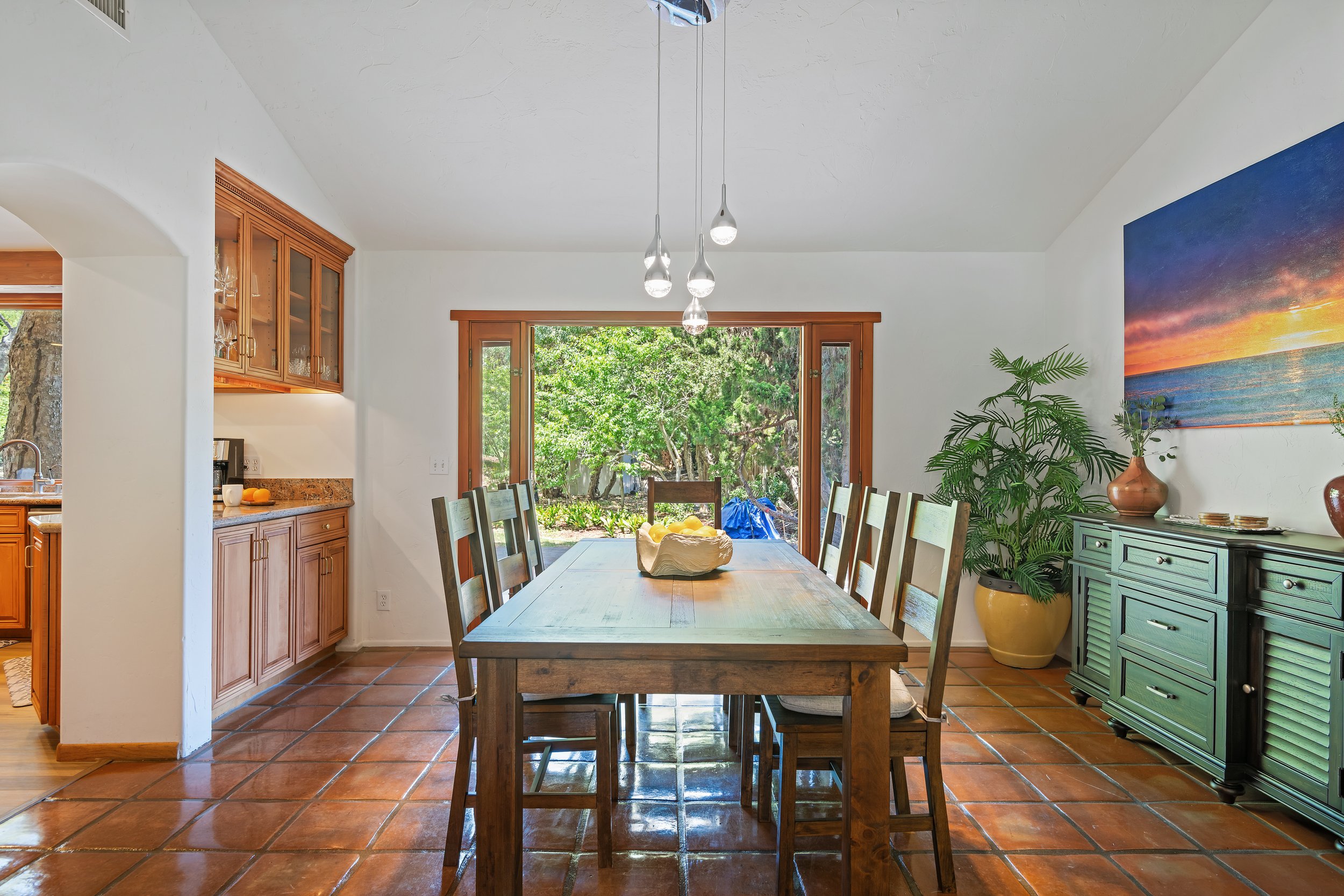 Dining room with wooden table and chairs, sliding glass door opening to yard, green cabinet, large potted plant, sunset painting, and terracotta tiled floor.