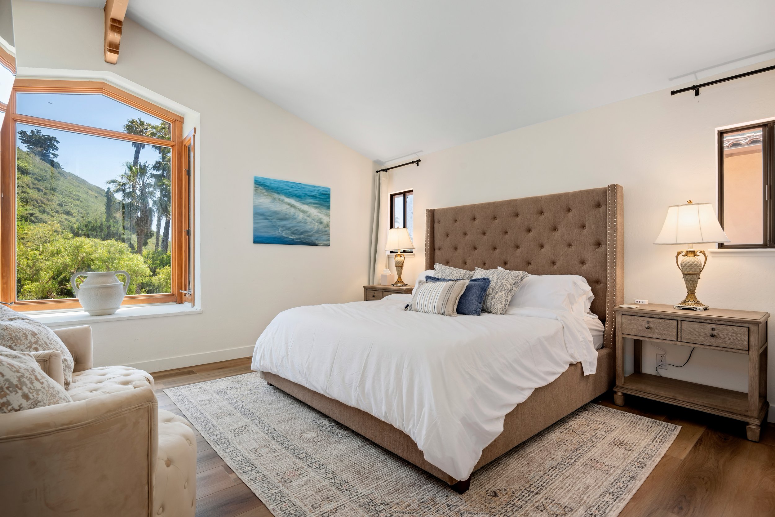 A bedroom with a large bed featuring a tan upholstered headboard, nightstands with lamps, a window showing a view of trees and hills, and a cream-colored armchair with pillows