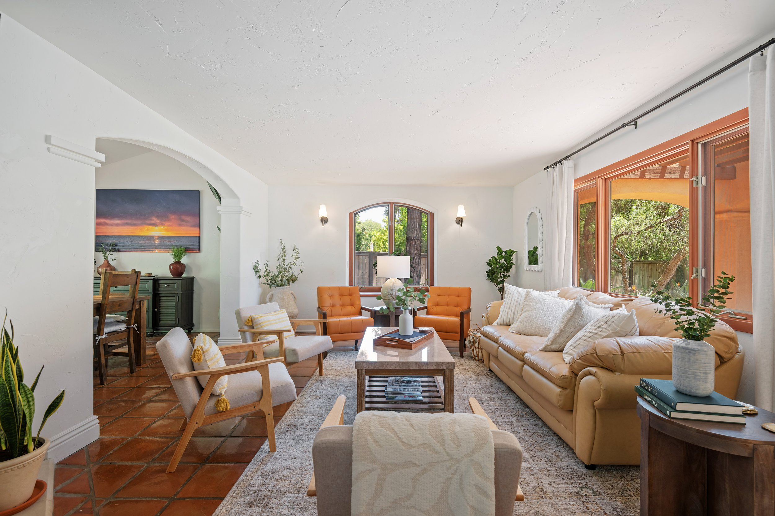 Living room with a large beige leather couch, orange chairs, and a wooden coffee table, decorated with pillows and plants, with large windows showing trees outside.