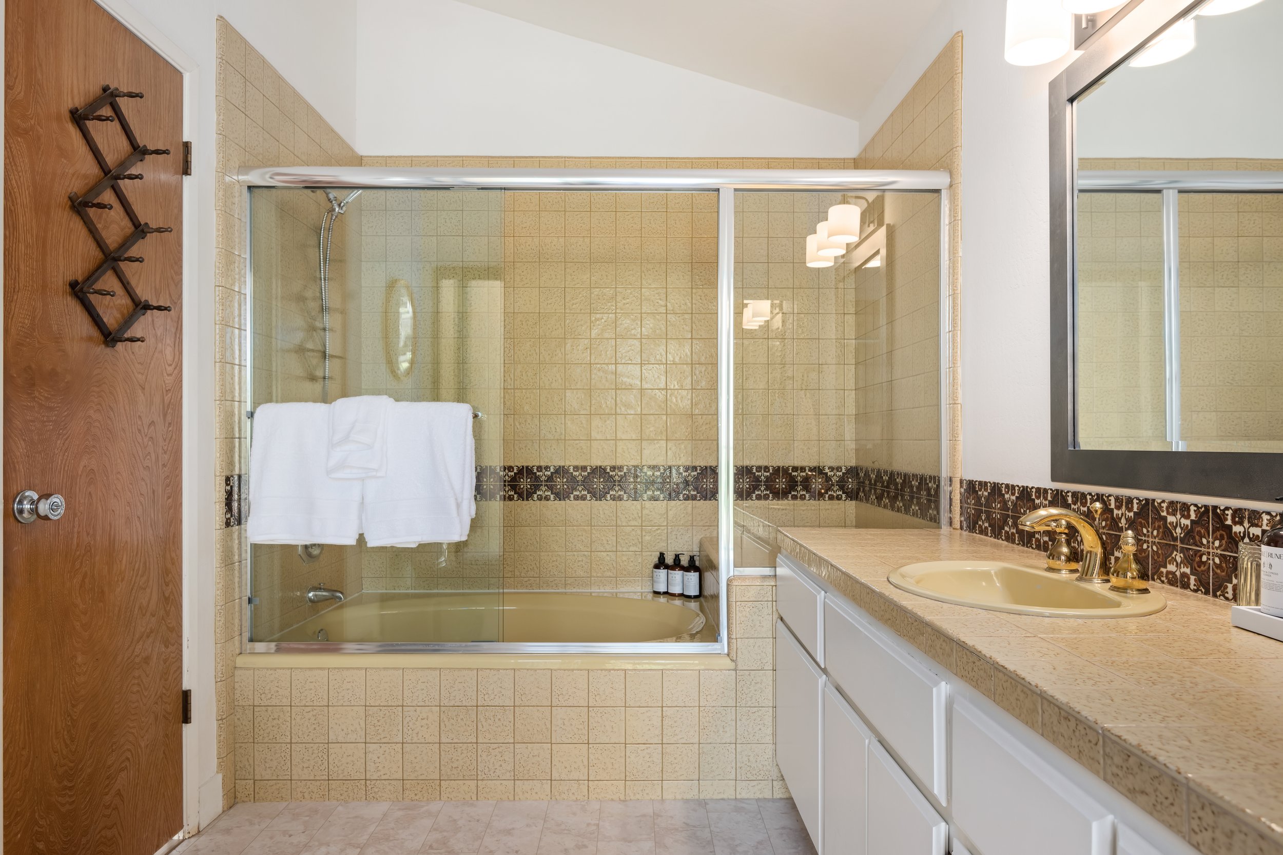 Bathroom with a beige tile bathtub and a gold faucet, beige countertop with a gold faucet, and a large mirror.