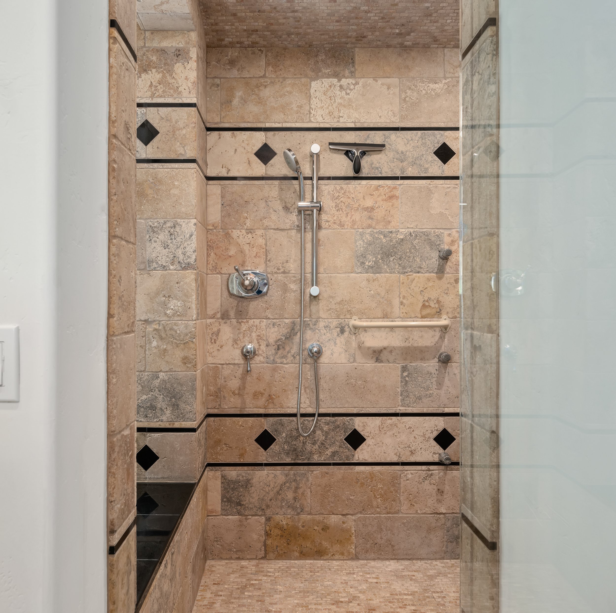 Walk-in shower with beige and brown stone tiles, black diamond accents, and a glass door.