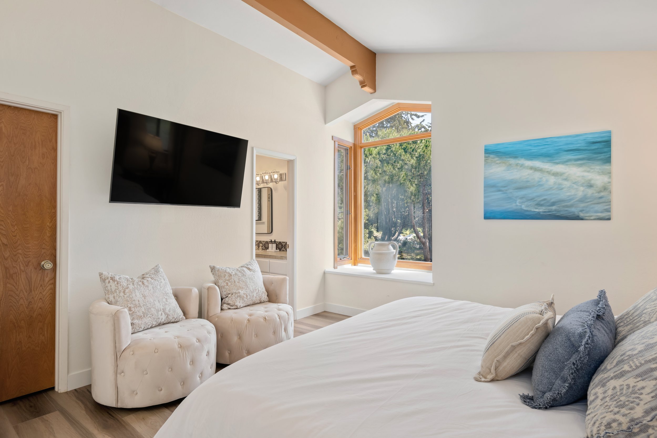 A bedroom with a large bed, a window with a vase on the sill, a wall-mounted flat screen TV, two tufted armchairs with patterned pillows, and a beach-themed painting on the wall.