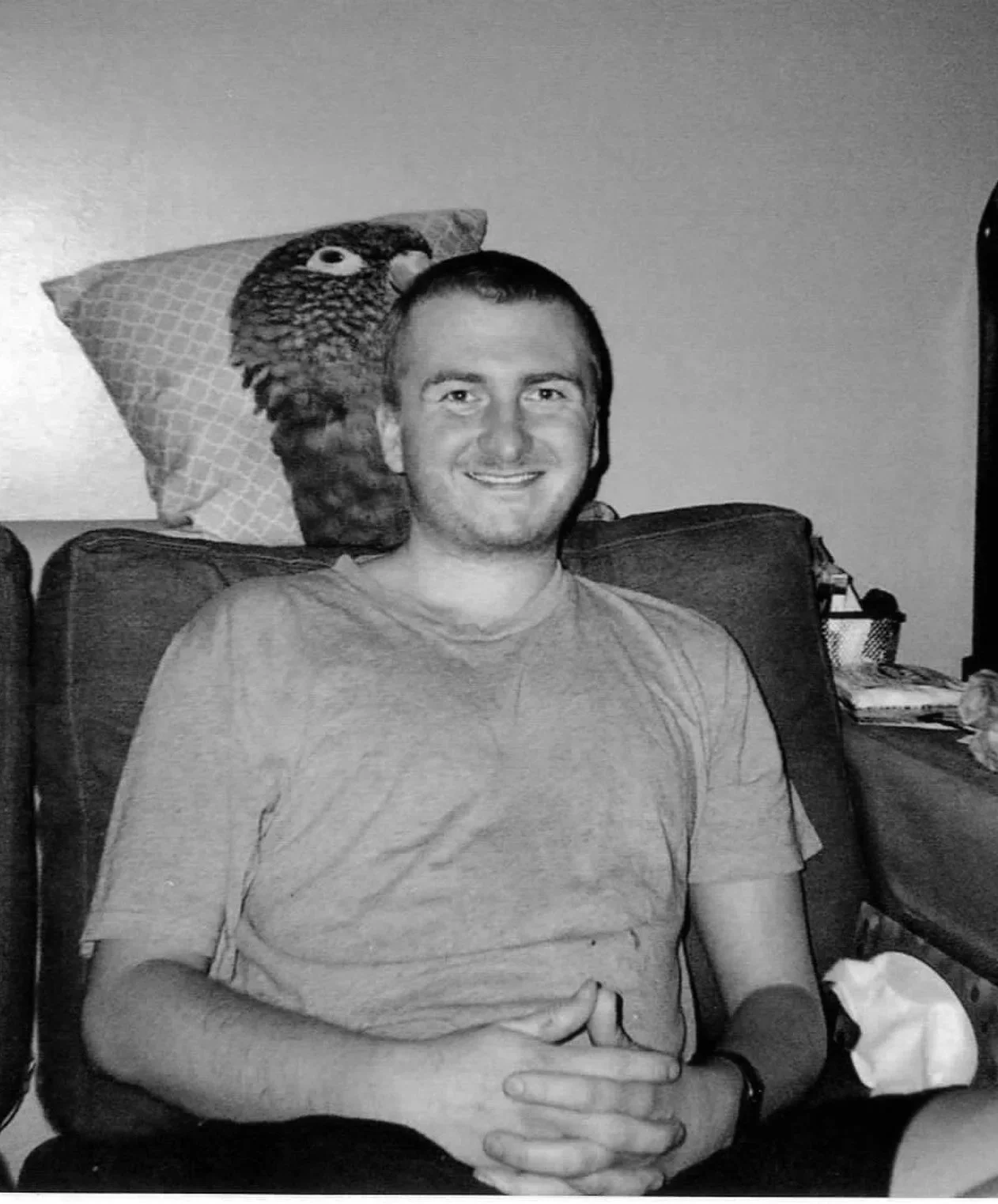 CJ is sitting relaxed in an armchair with his hands clapsed on his lap. His has short hair and is smiling. There is a noticable cushion with a parrot on behind his head.