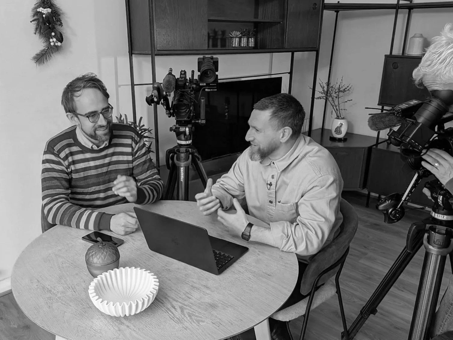 Matt and the presenter sat at a table having a conversation. Around the table are studio cameras