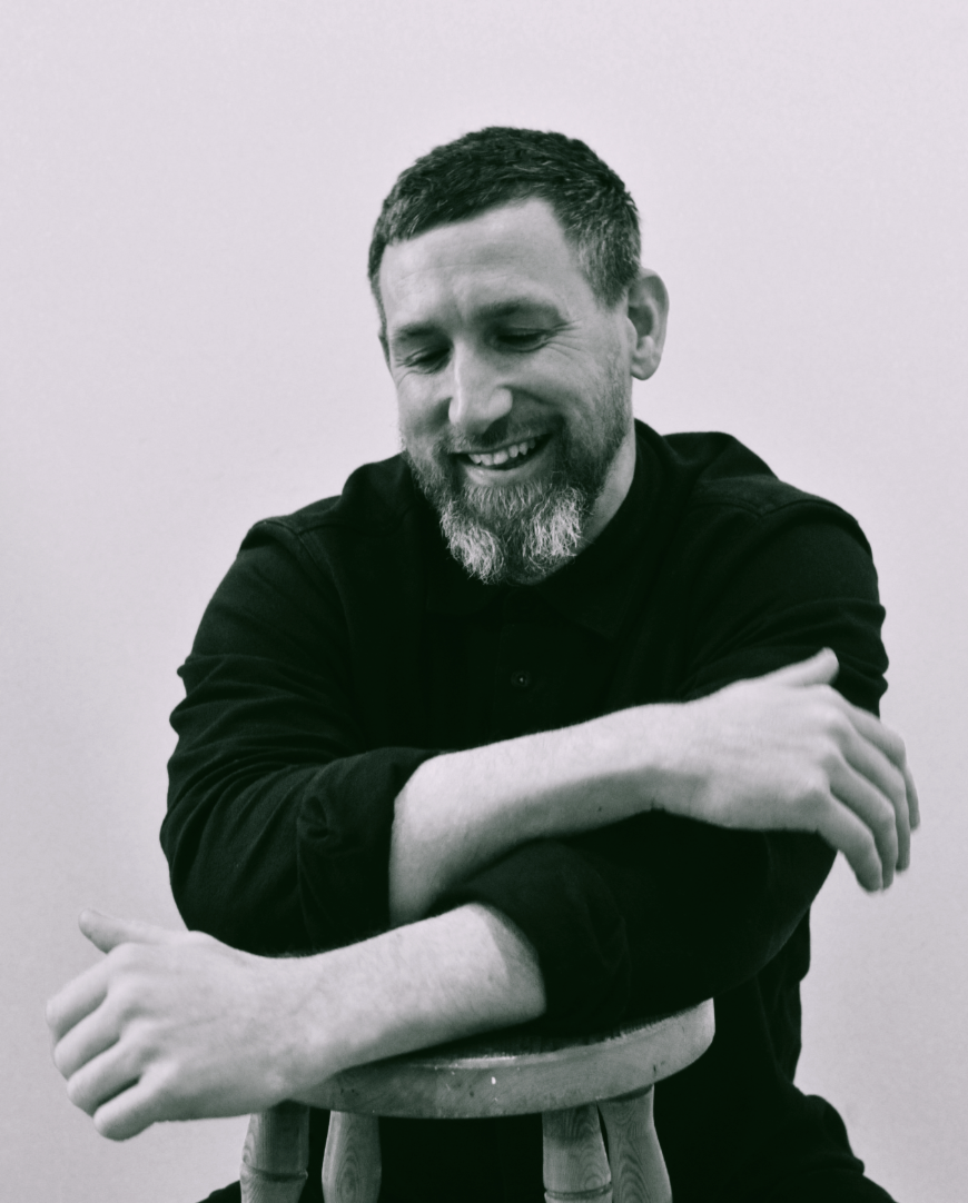 Matt is smiling whilst leaning his loosely crossed arms on a stall. His greying beard is quite something.