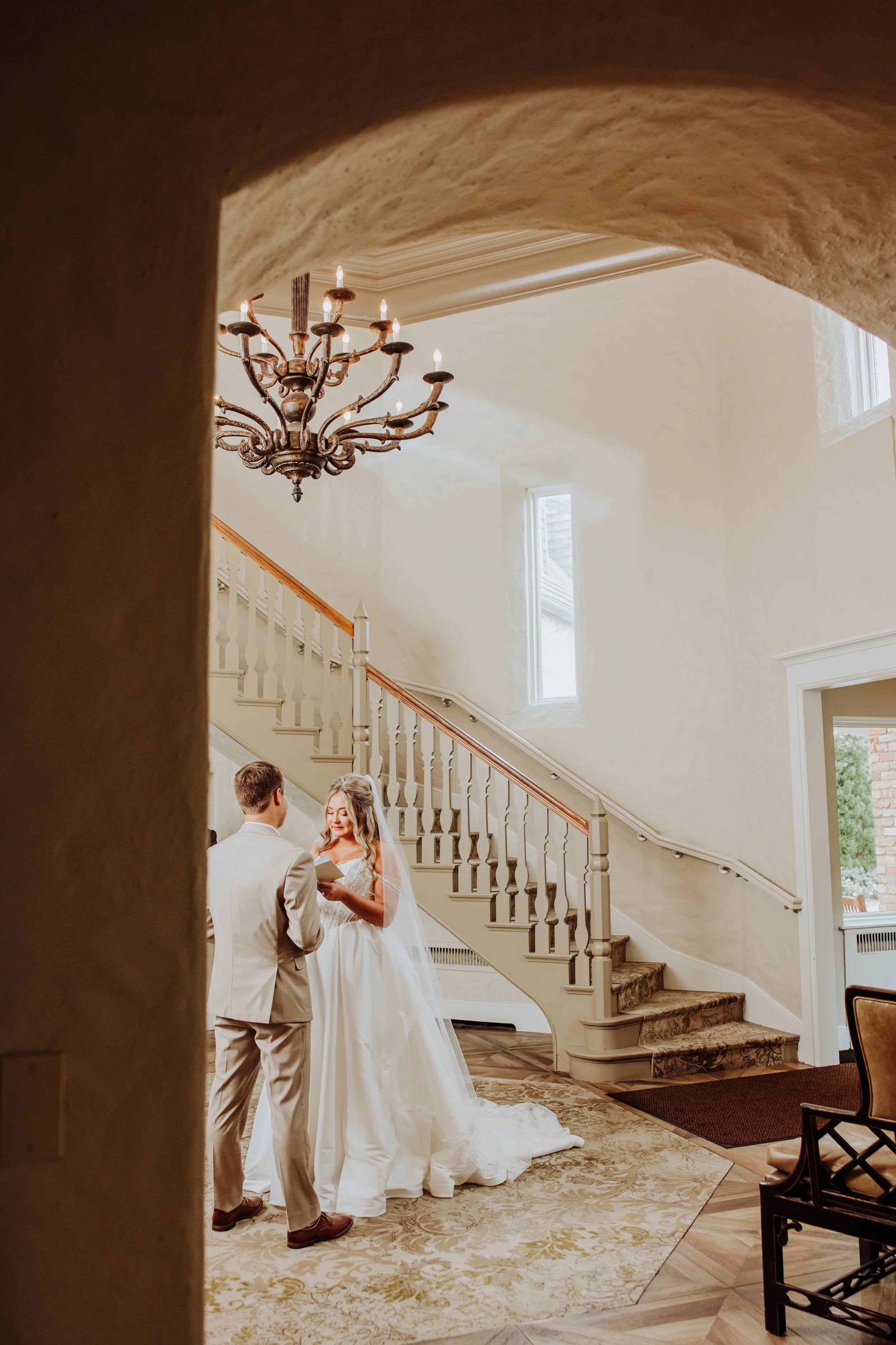 A bride and groom standing together inside a well-lit foyer with high ceilings and a staircase. The bride is in a white wedding dress, and the groom is in a beige suit. They are exchanging vows or rings, with a chandelier hanging above inside the hou