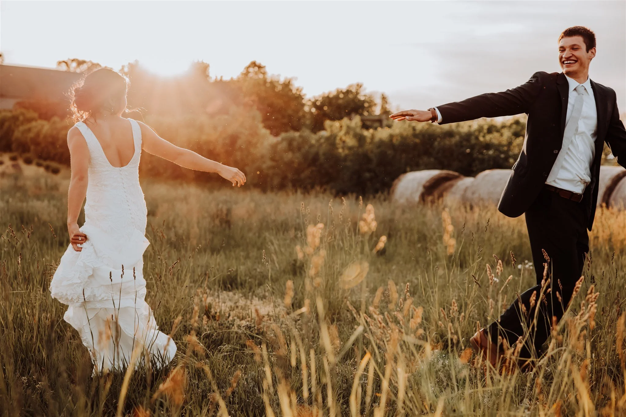 A bride and groom holding hands in a field during sunset, both smiling joyfully.