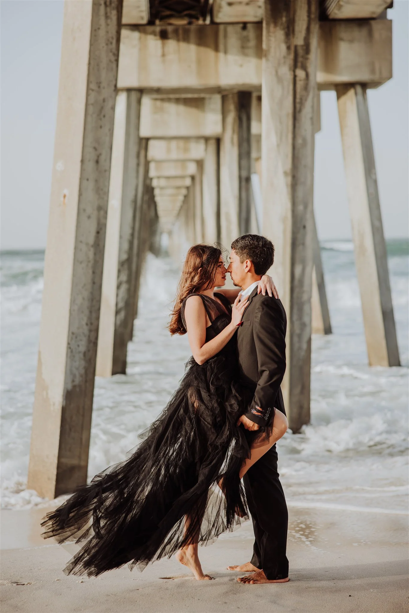 A couple stands close together on the beach under a pier, with the woman in a black dress and the man in a black suit, both barefoot, their faces close, about to kiss.