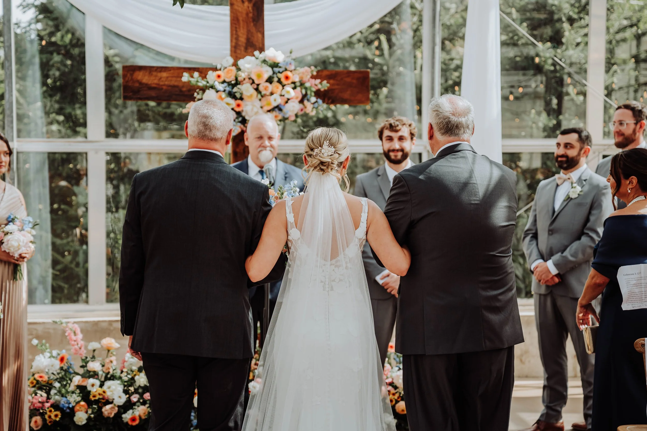 A bride and groom standing together during their wedding ceremony, surrounded by friends and family, with an outdoor floral and cross altar behind them.