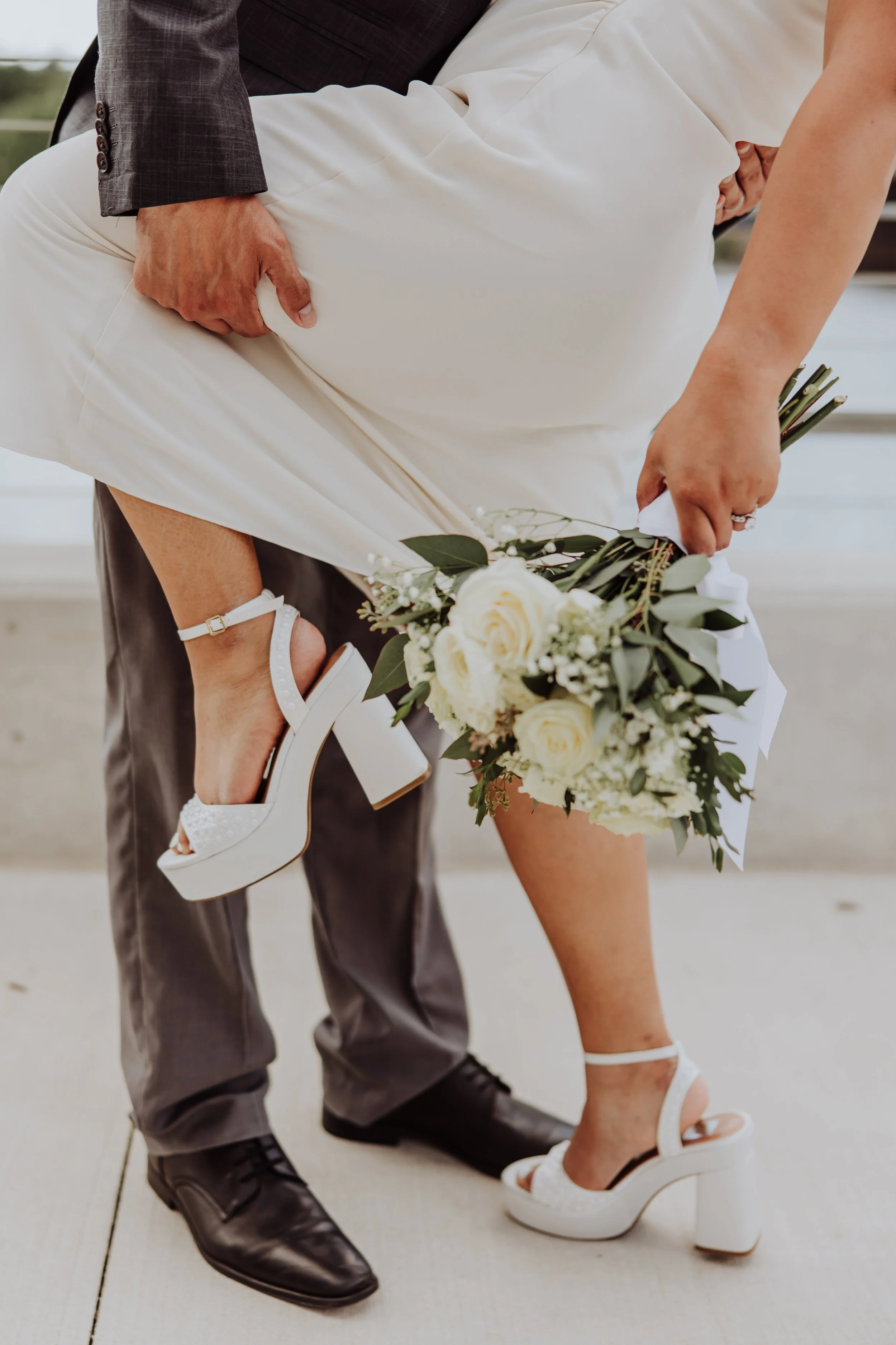 Close-up of a couple on their wedding day, with the bride holding a bouquet of white roses and greenery, and the groom holding her leg as she stands in white high-heeled sandals, while he is in black formal shoes.