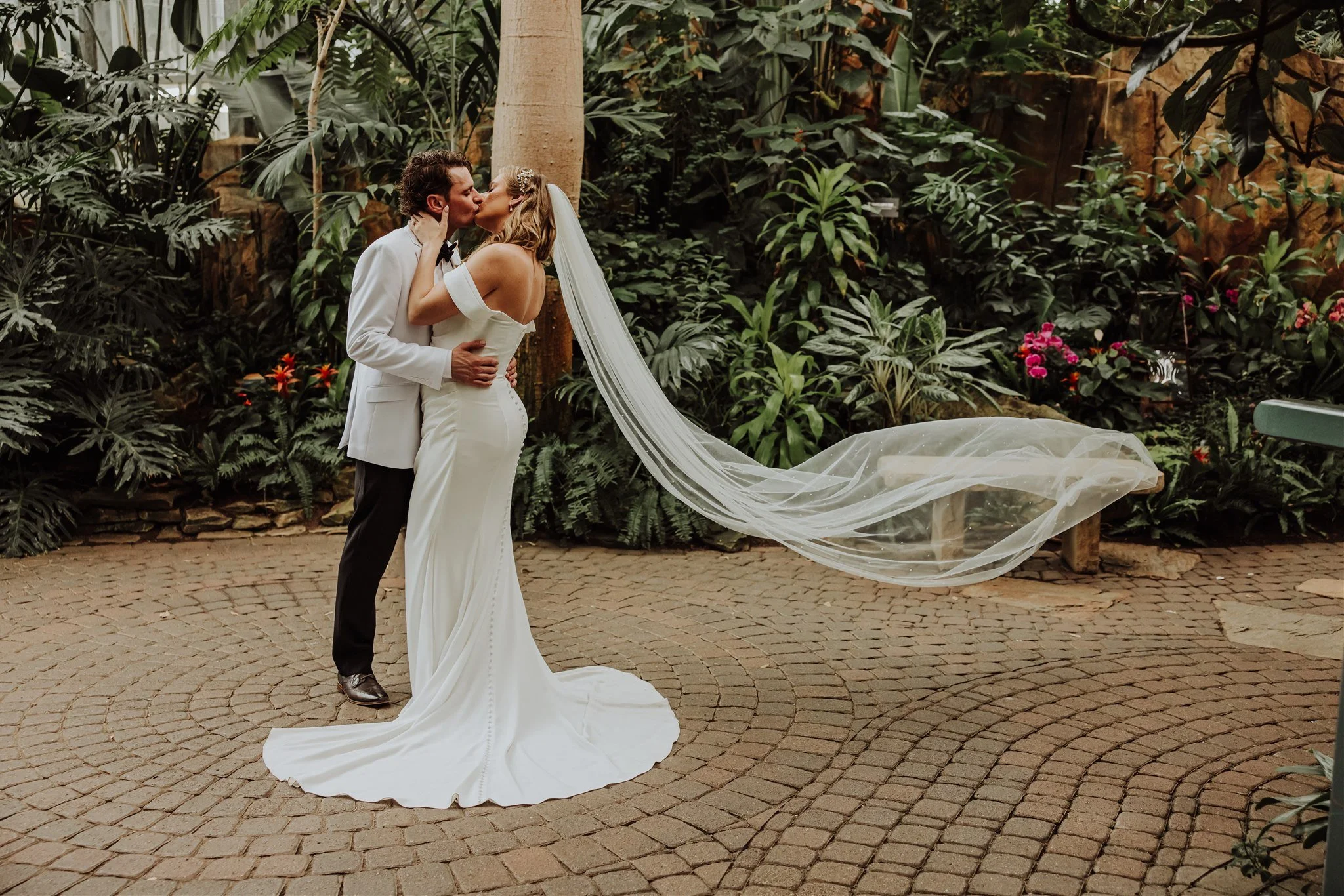 A bride and groom sharing a kiss in a lush indoor botanical garden, with the bride in a white off-shoulder gown and veil, and the groom in a white tuxedo jacket and black pants, standing on a cobblestone path surrounded by large green plants and flow