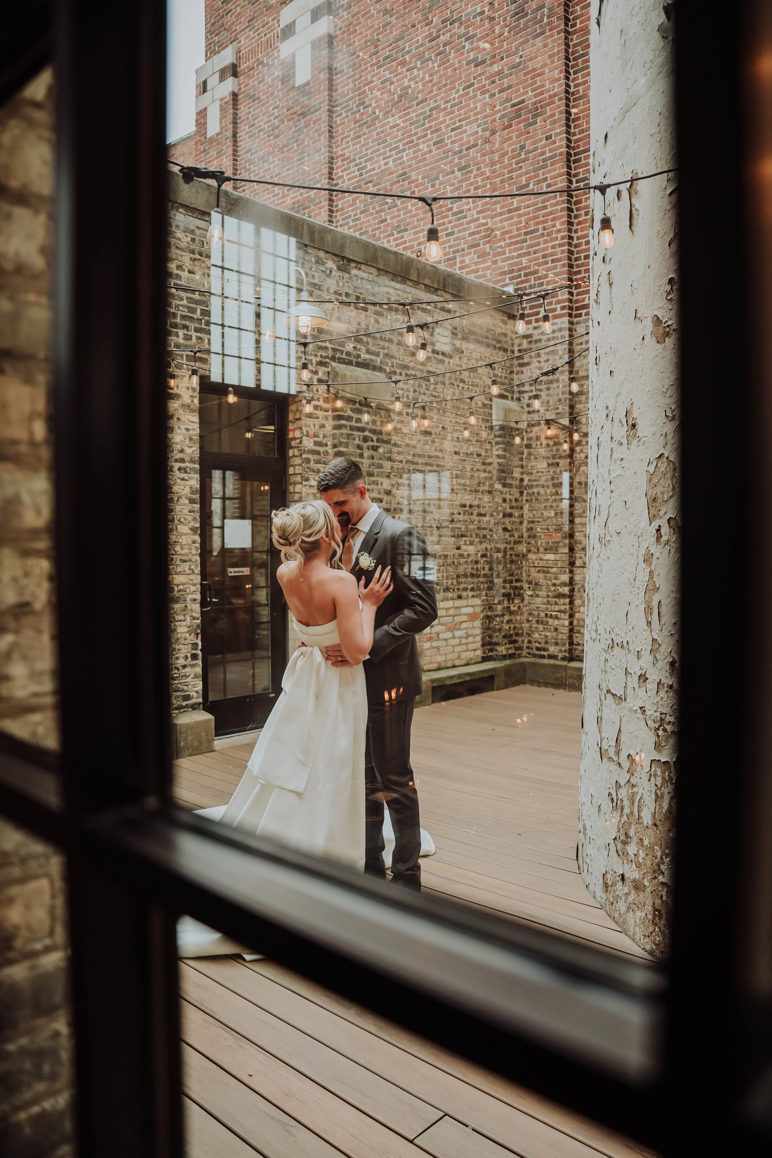 A bride and groom dancing during their wedding reception in an urban outdoor setting with brick walls and string lights, viewed through a window.