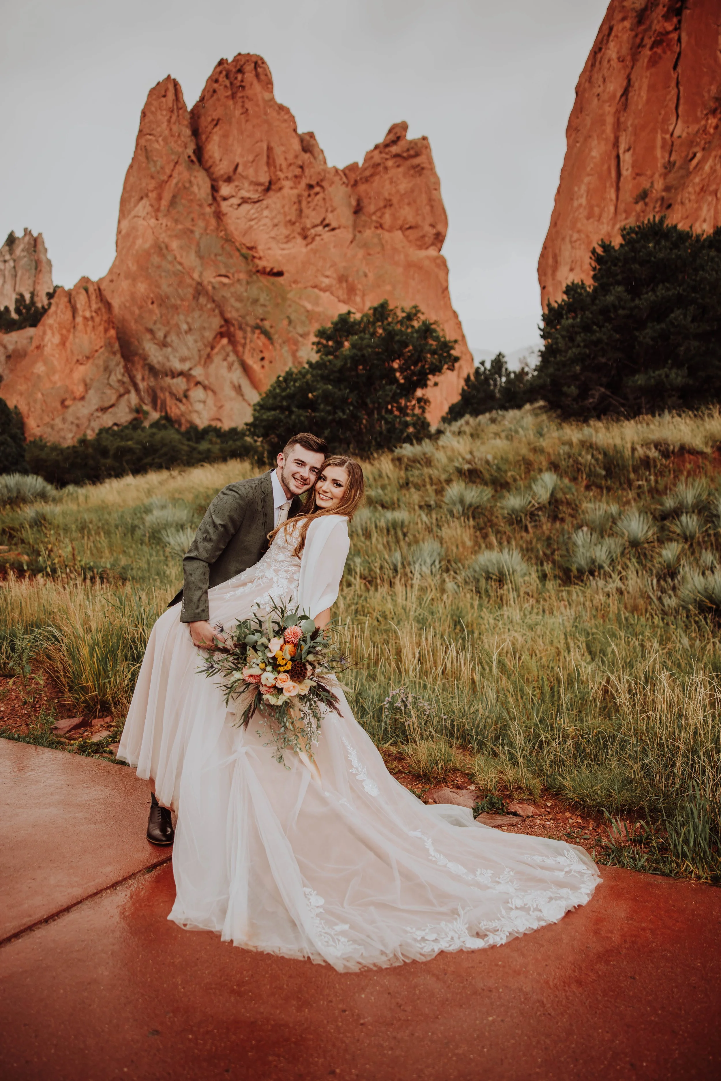 A newlywed couple in wedding attire smiling and embracing outdoors in front of red rocky cliffs and green trees.