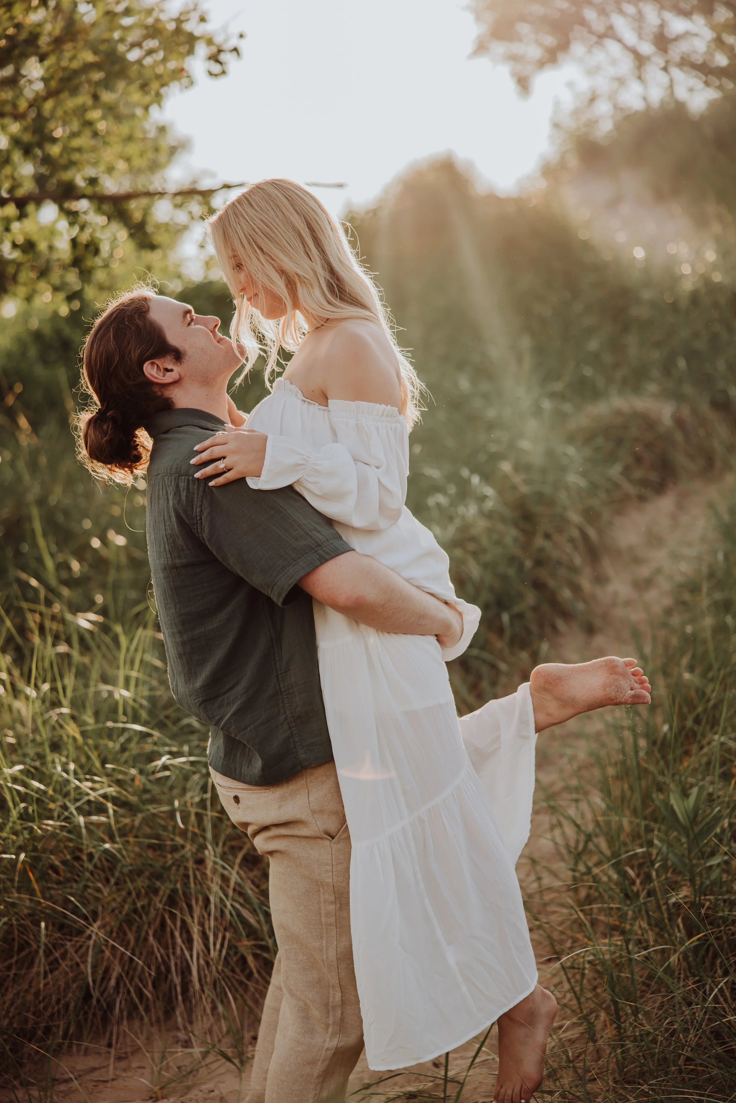 A happy couple outdoors, the man lifting the woman, both smiling and looking into each other's eyes, with greenery and sunlight filtering through trees in the background.