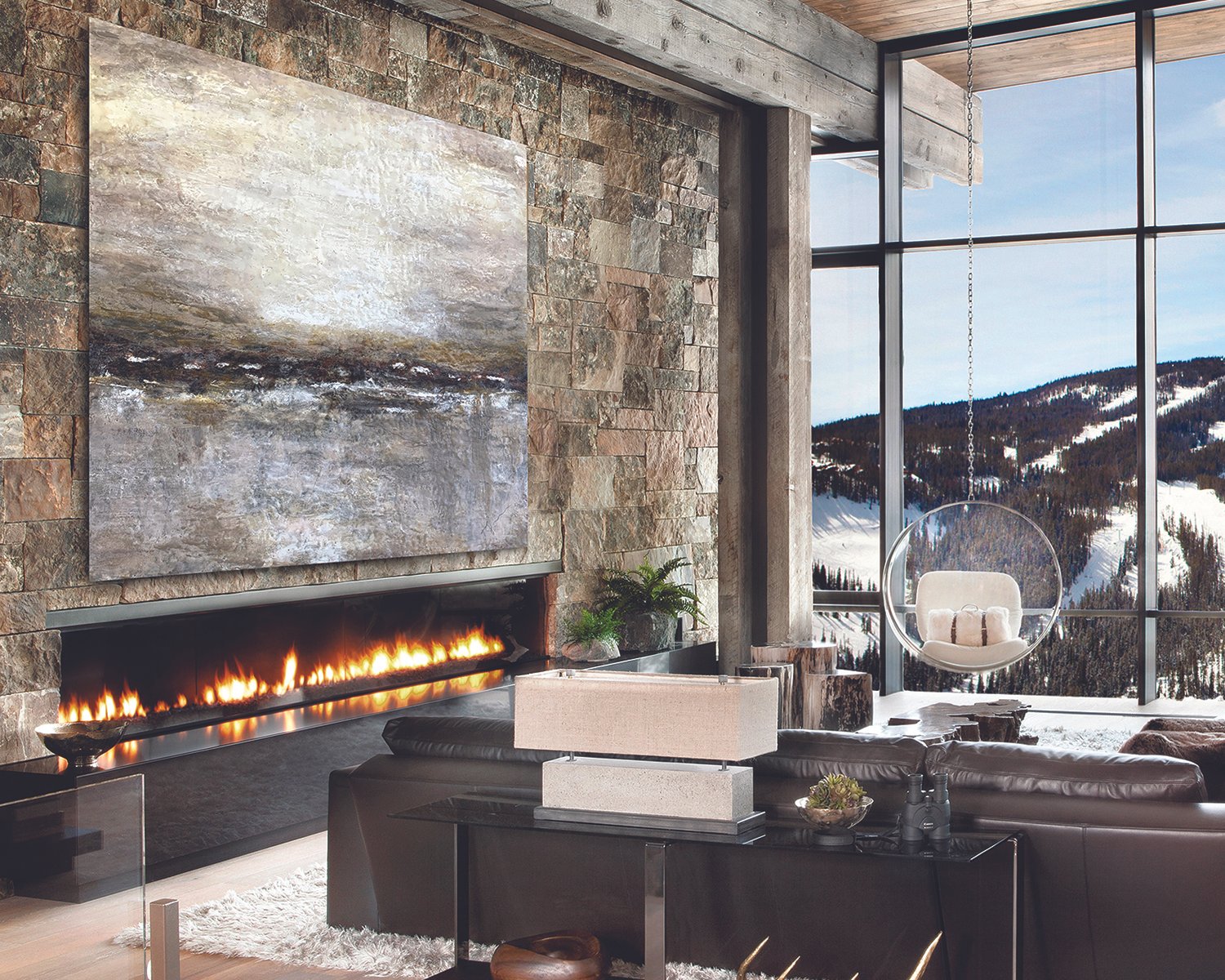 Modern living room with a large stone wall, a fireplace with a linear fire, a current abstract painting, a black leather sofa, a small table with binoculars, a white hanging bubble chair, and large windows showing snowy mountains and trees.