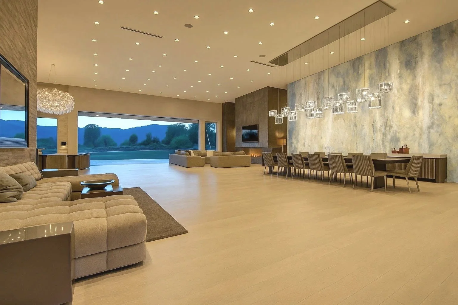 Spacious modern living room with beige sofas, large windows overlooking mountains, and elegant dining area with glass chandelier.
