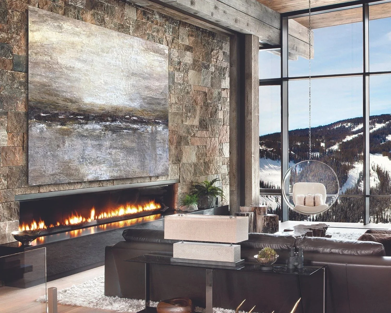 Modern living room with a large stone wall, a fireplace with a linear fire, a current abstract painting, a black leather sofa, a small table with binoculars, a white hanging bubble chair, and large windows showing snowy mountains and trees.
