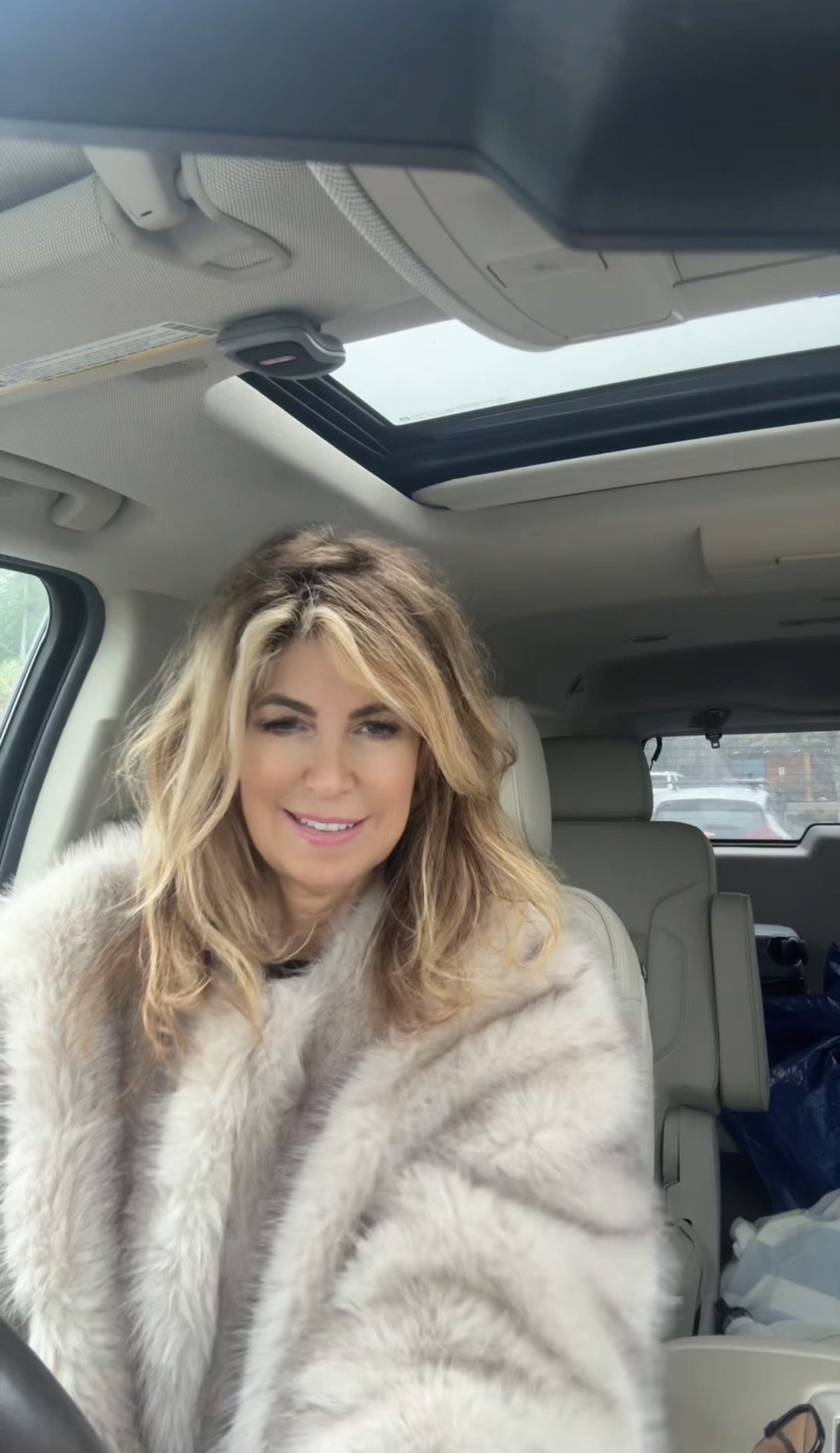 A woman with blonde, wavy hair smiling inside a vehicle with a sunroof and beige interior.