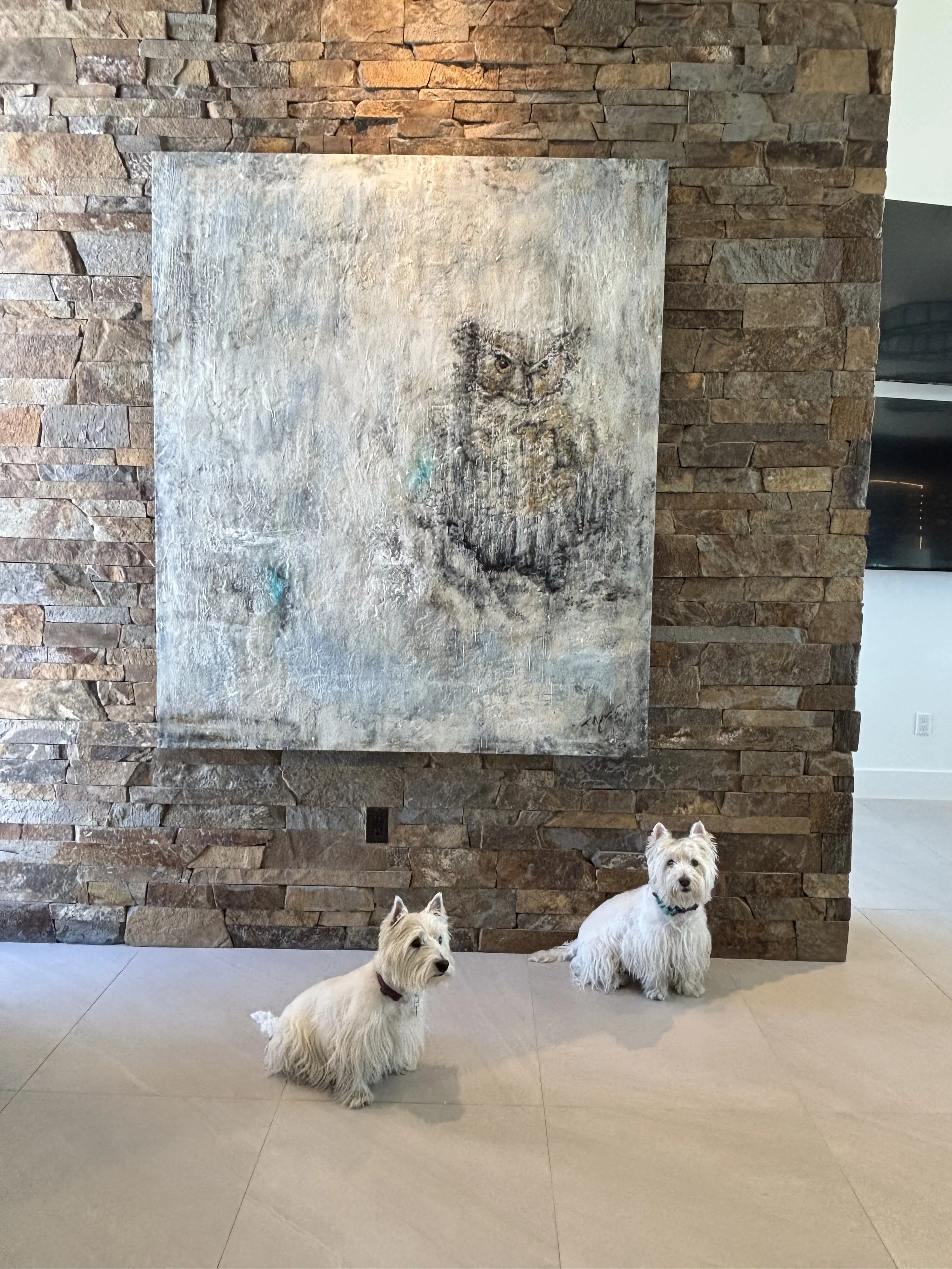Two small white dogs sitting on beige tile floor in front of a stone wall, with a large abstract painting of an owl hanging on the wall behind them.