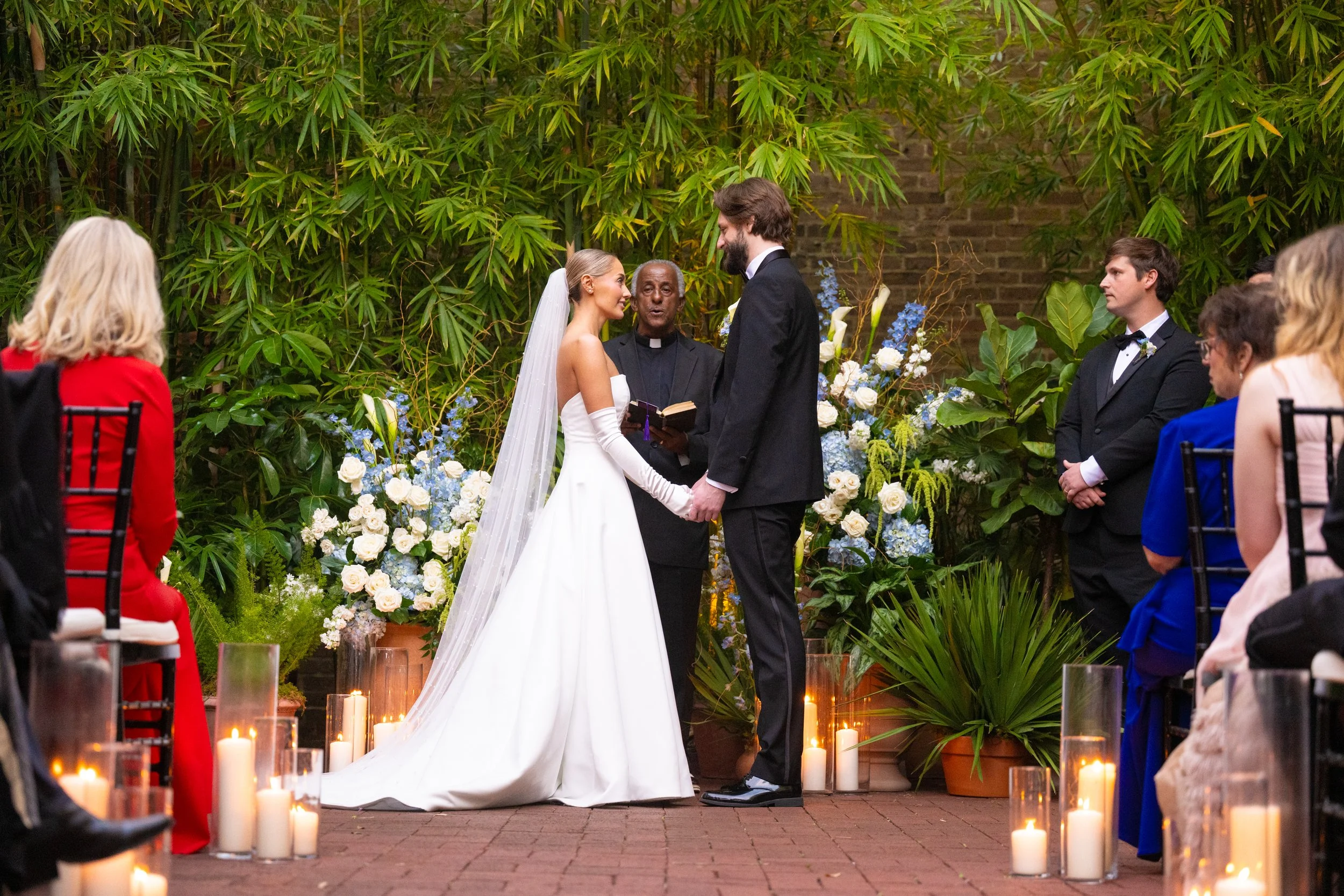 Bride and groom exchanging vows during outdoor wedding ceremony, surrounded by green foliage and candles.