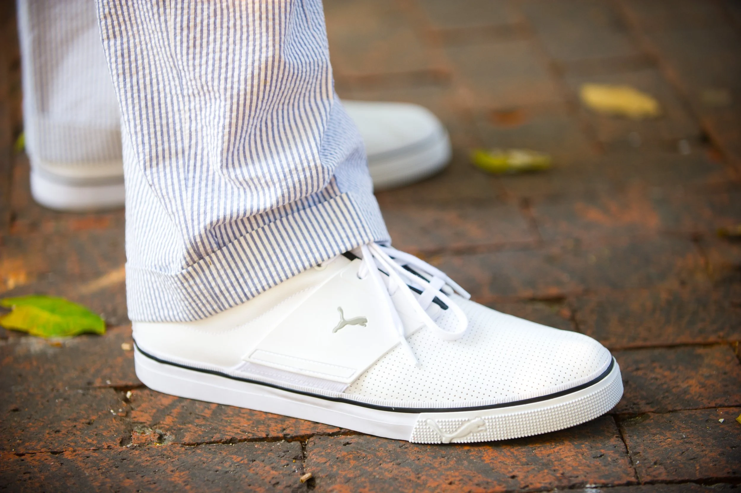 Close-up of a person wearing white Puma sneakers and light blue and white striped pants, standing on a wet brick sidewalk with fallen leaves.