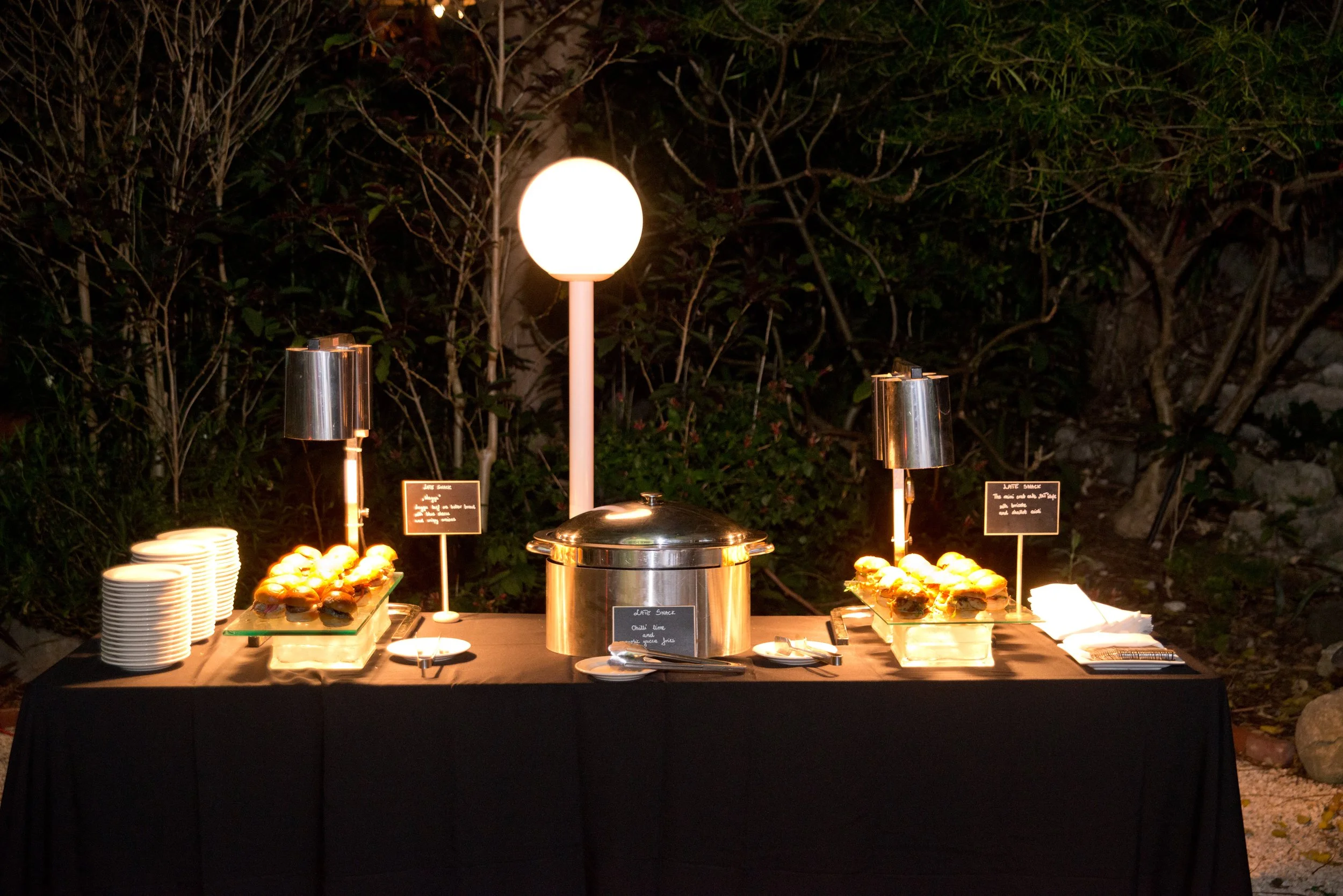 An outdoor buffet table set up at night with various foods, including snacks, on a table with black tablecloth. There are lights and small signs identifying the food items, with a background of bushes and trees.