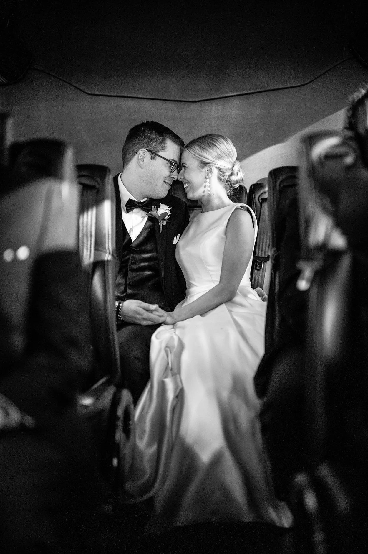 A black and white photo of a bride and groom sitting together inside a vehicle, touching foreheads and smiling at each other, holding hands.