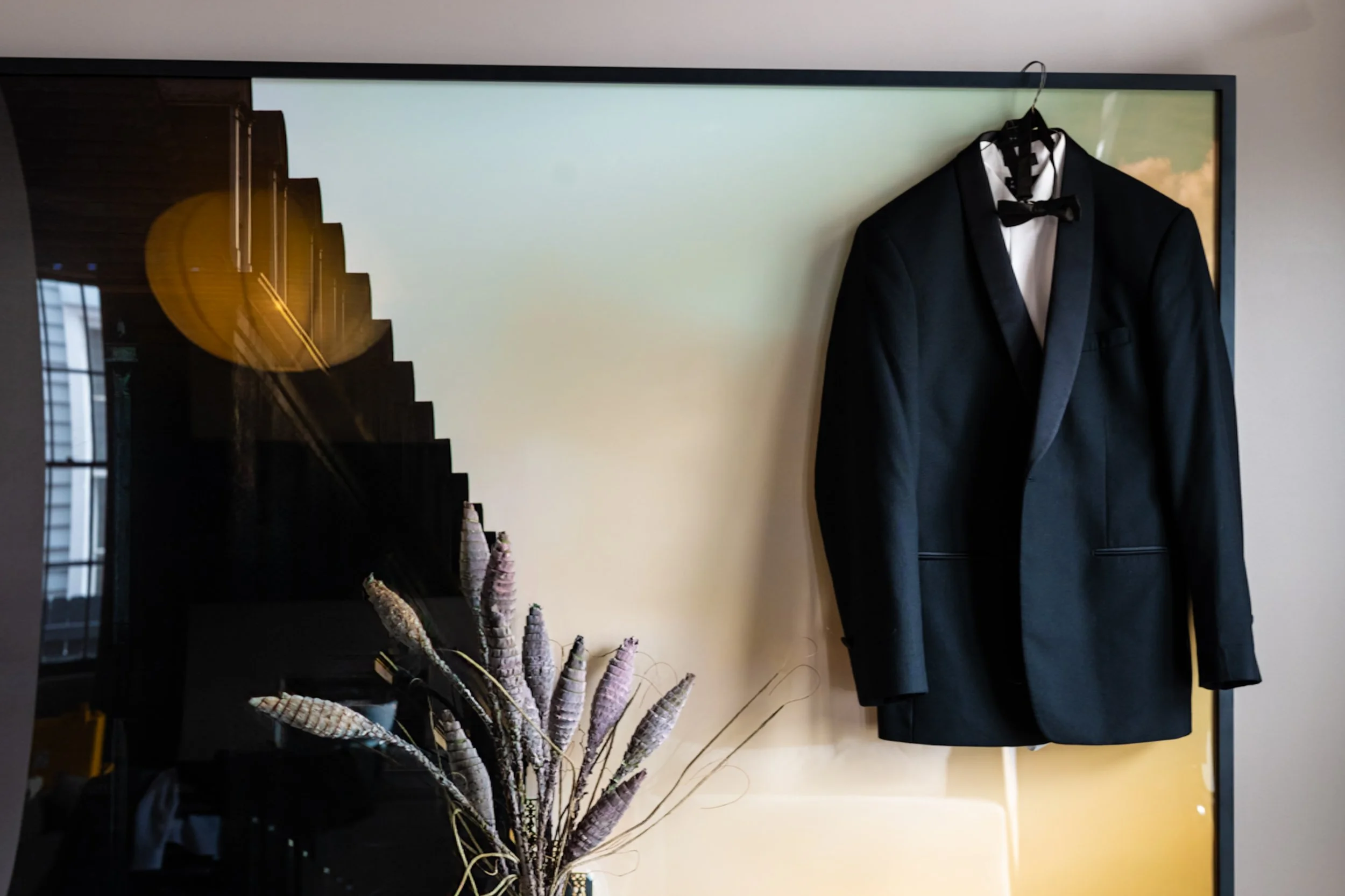 A black tuxedo jacket with a white shirt and black bowtie hanging on a hook on the wall. A decorative plant with tall, spiked, purplish stalks is in the foreground, and a window with reflections is visible on the left side of the image.